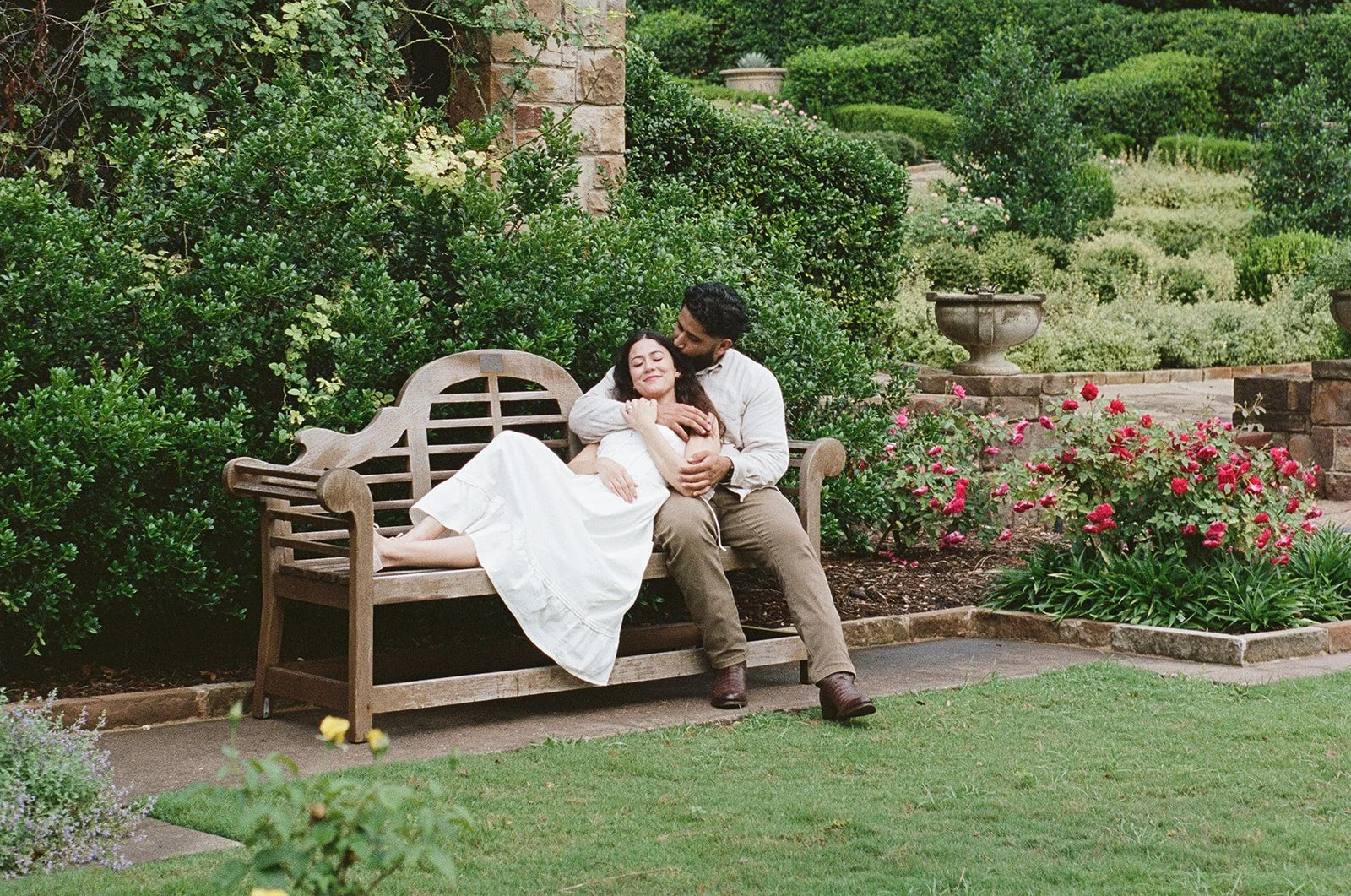 A couple sitting on a wooden bench in a garden surrounded by green bushes and colorful flowers. The woman is smiling with her eyes closed while the man is lovingly embracing her.