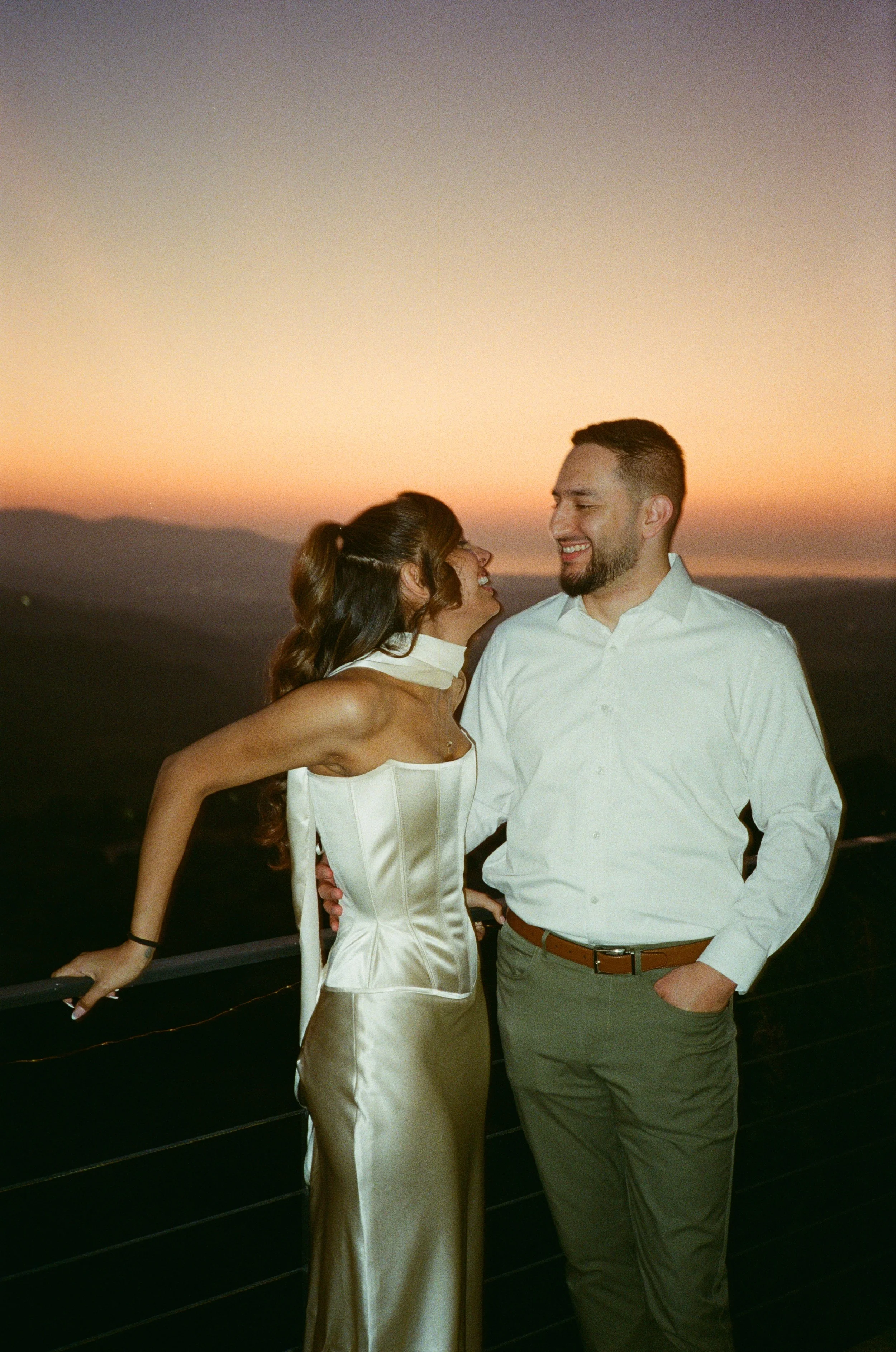 A couple smiling and looking at each other at sunset, with rolling hills and mountains in the background.