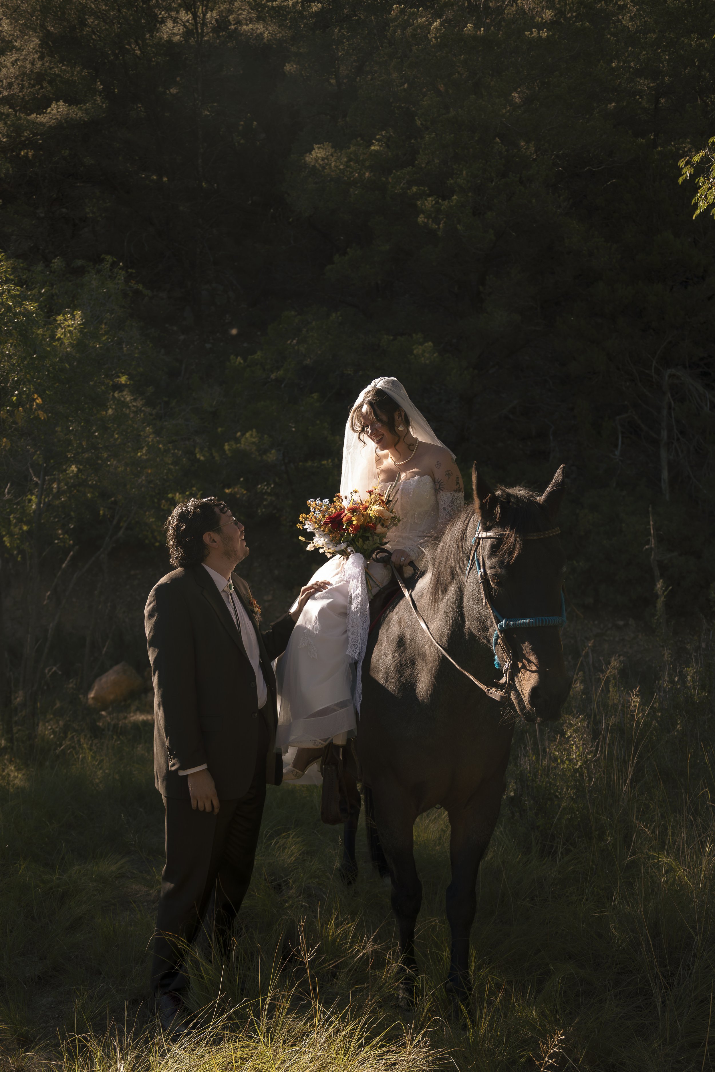 A bride in a white wedding dress and veil sitting on a horse, holding a bouquet of flowers, with a man in a suit standing beside her, in a natural outdoor setting with trees and grass.