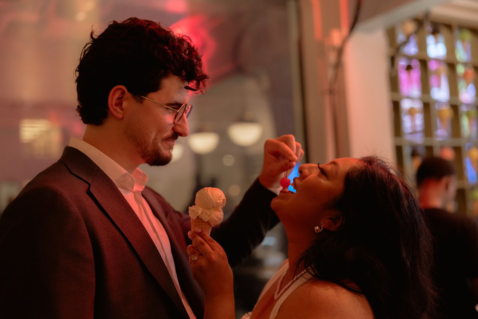 A man in a suit and glasses is holding an ice cream cone while a woman with earrings is feeding him a cherry from a spoon inside a colorful restaurant or cafe.
