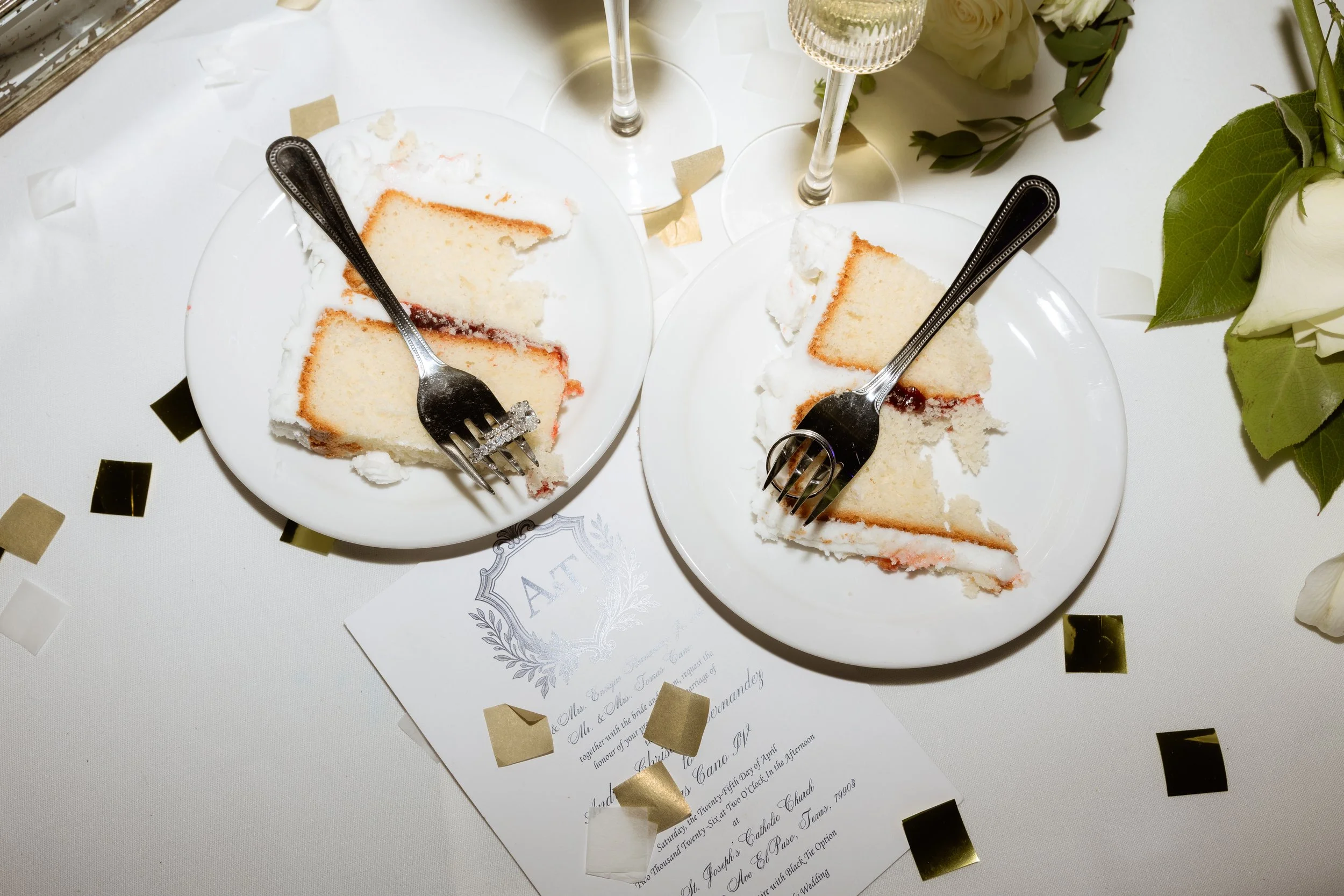 Two plates with slices of white cake with strawberry jam and whipped cream, forks on the plates, two champagne flutes, white flowers, portion of wedding invitation card, and gold confetti on a table.