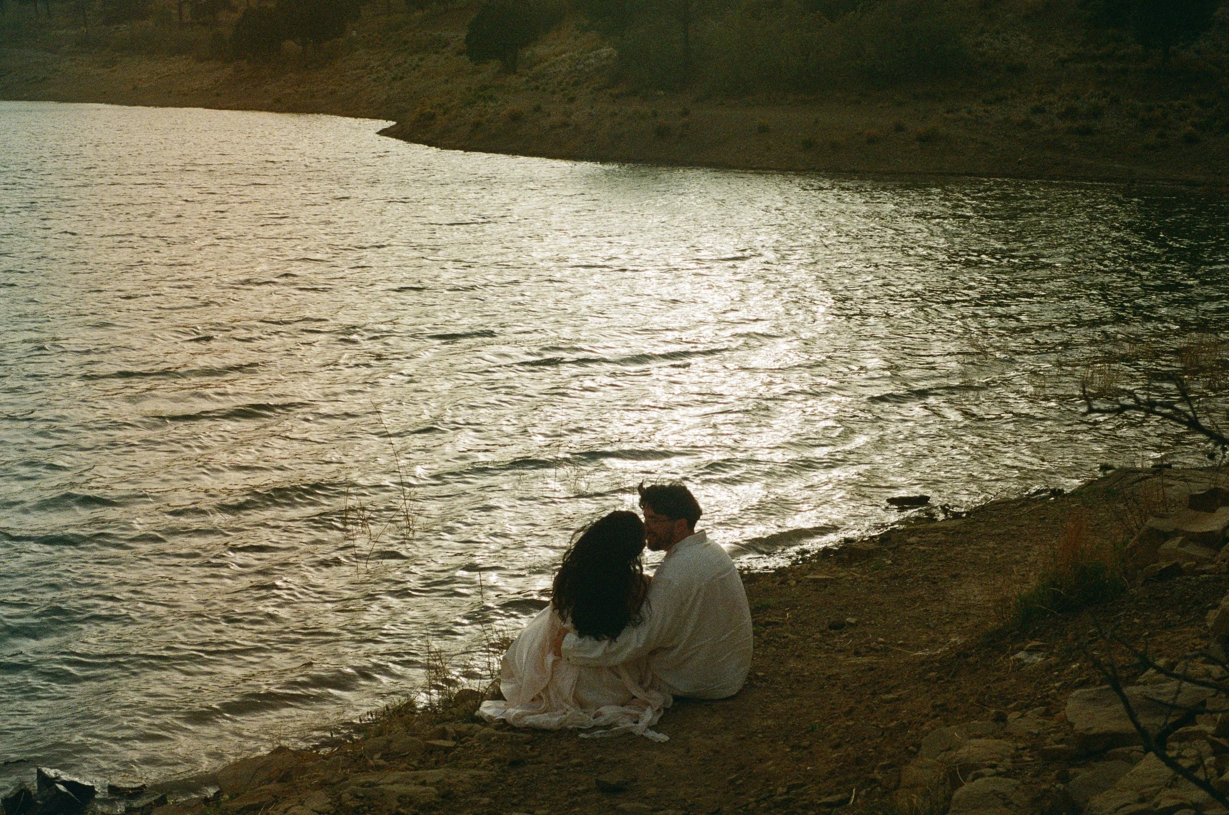 A couple sitting by the water's edge on a rocky shore, with a hilly landscape in the background. The sunset reflects on the water, creating a peaceful scene.