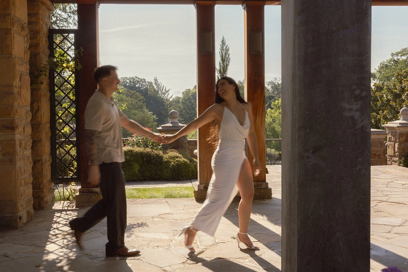 A man and woman dance together outdoors, holding hands and smiling, with a scenic background of trees and sky. The woman is wearing a white dress and high heels, and the man is dressed casually.
