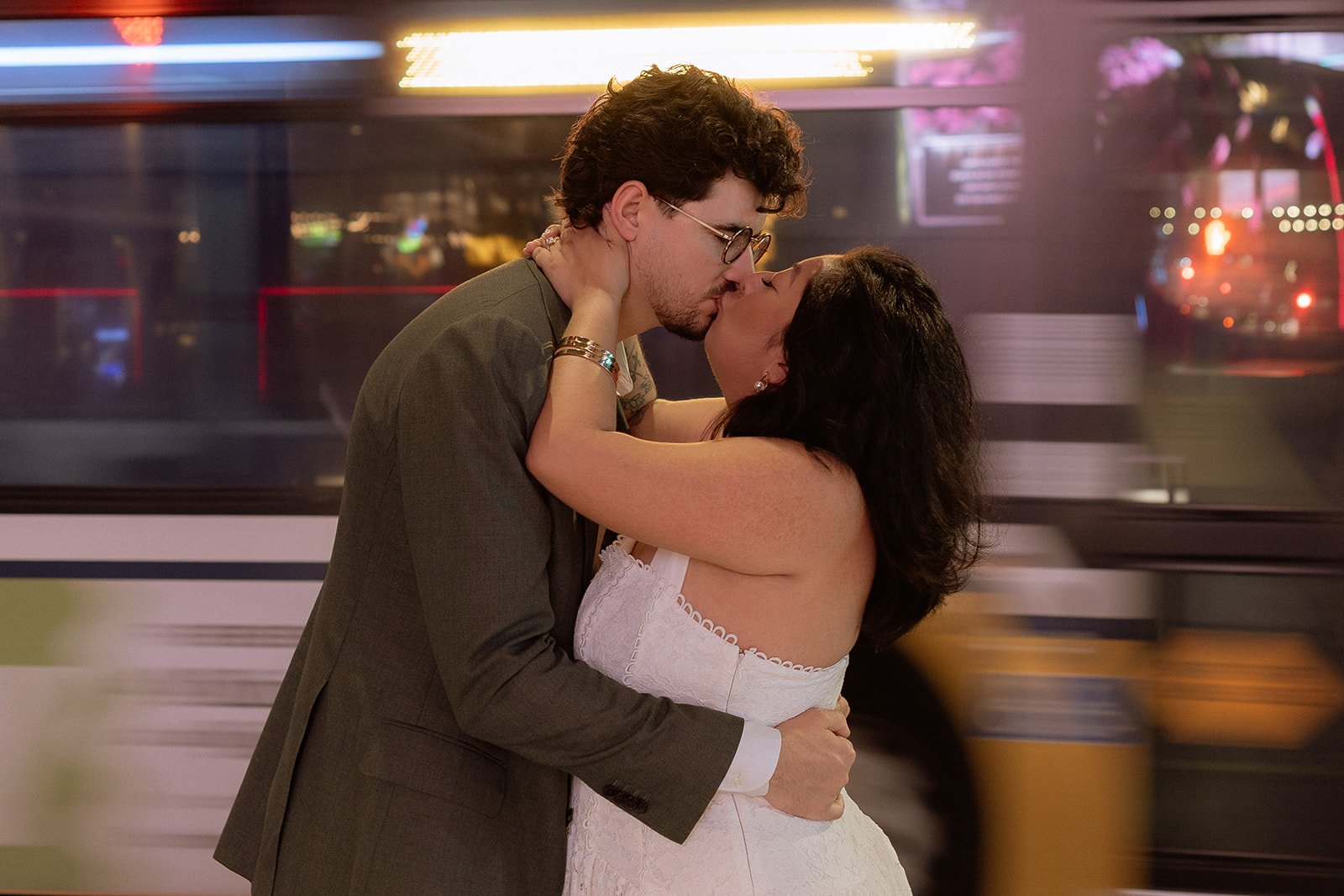 A couple sharing a kiss on a city street at night with a motion-blurred bus passing behind them.