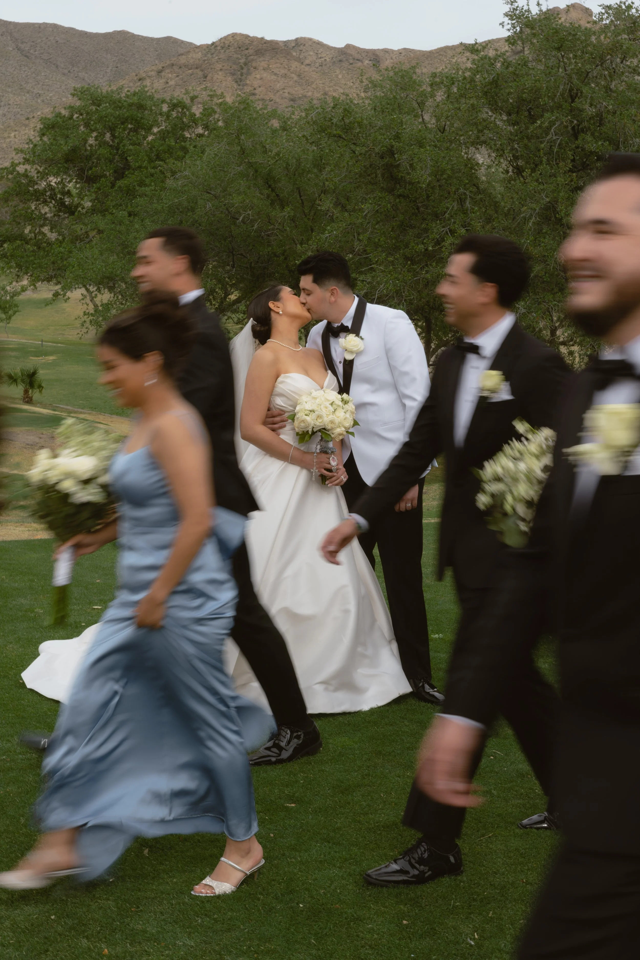 A wedding scene outdoors with the bride and groom kissing surrounded by dancing guests. The bride wears a white gown holding a bouquet, and the groom wears a white tuxedo with a black bow tie. Guests in formal attire walk by on a grassy area with gre