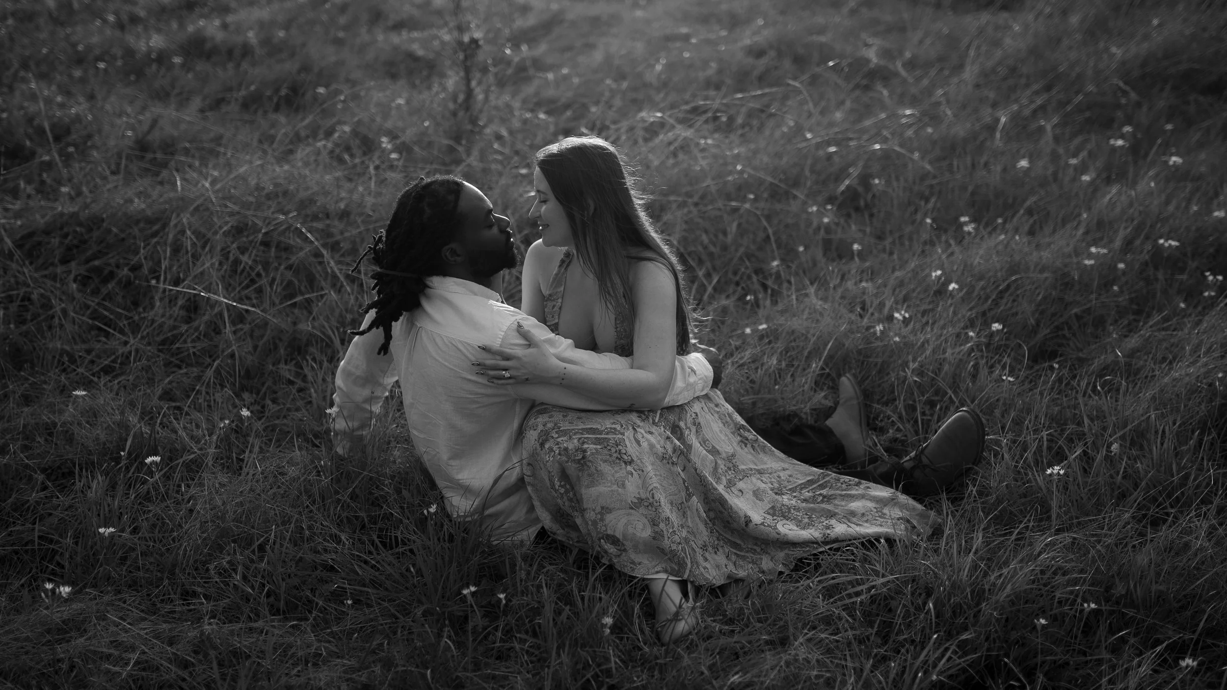 A black and white photo of a man and woman sitting close together in a grassy field, holding each other and smiling with their faces close.