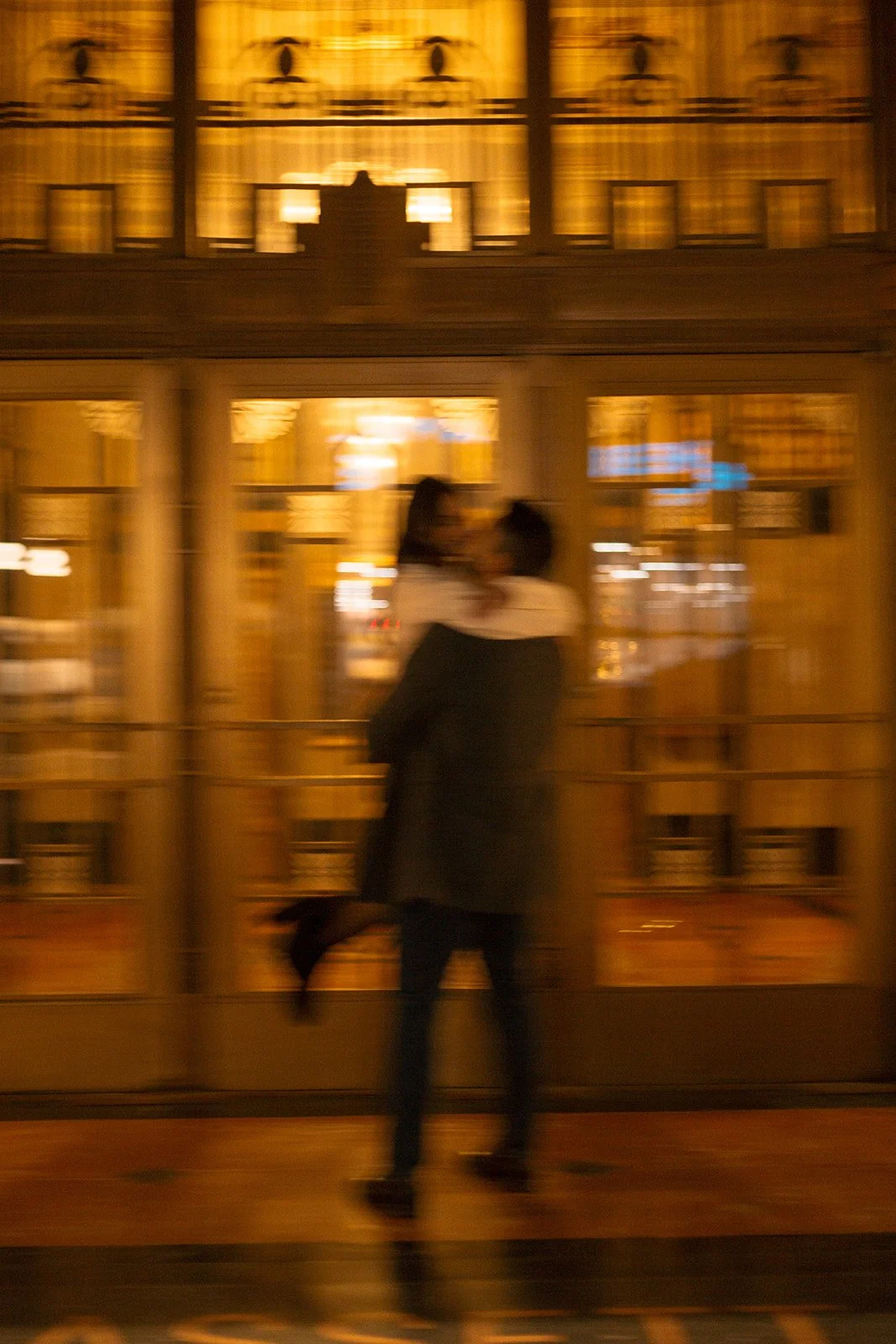 Two people embrace in front of a glass storefront with warm interior lighting, blurred and out of focus.
