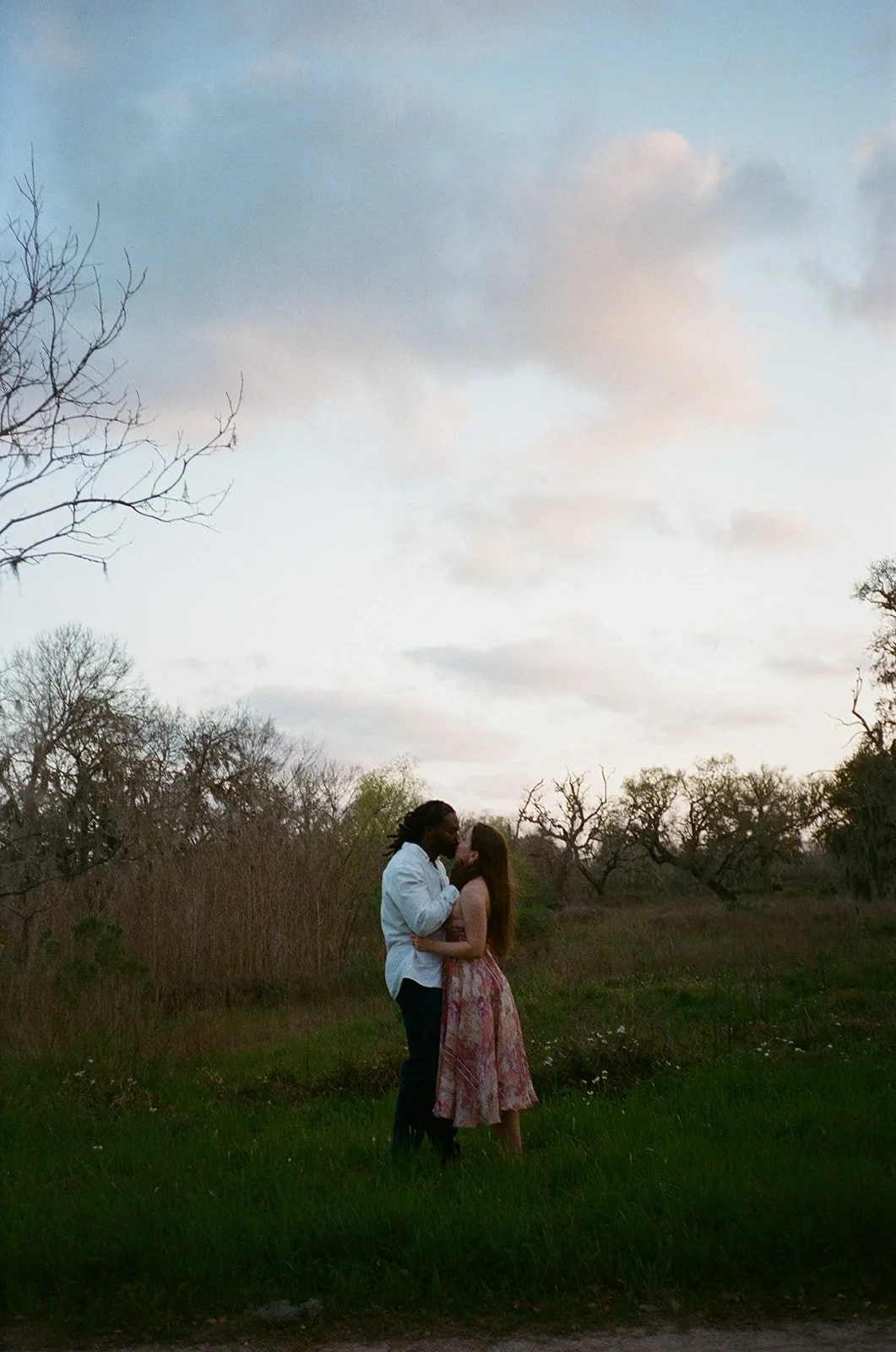 A couple kissing in a grassy field with trees and a cloudy sky in the background.