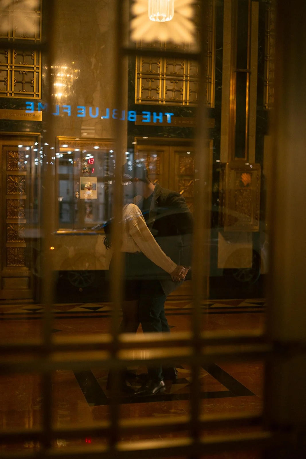 People holding hands in front of a window inside a luxurious, gold-decorated hotel lobby, with the sign 'The Bluebird' visible through the window.