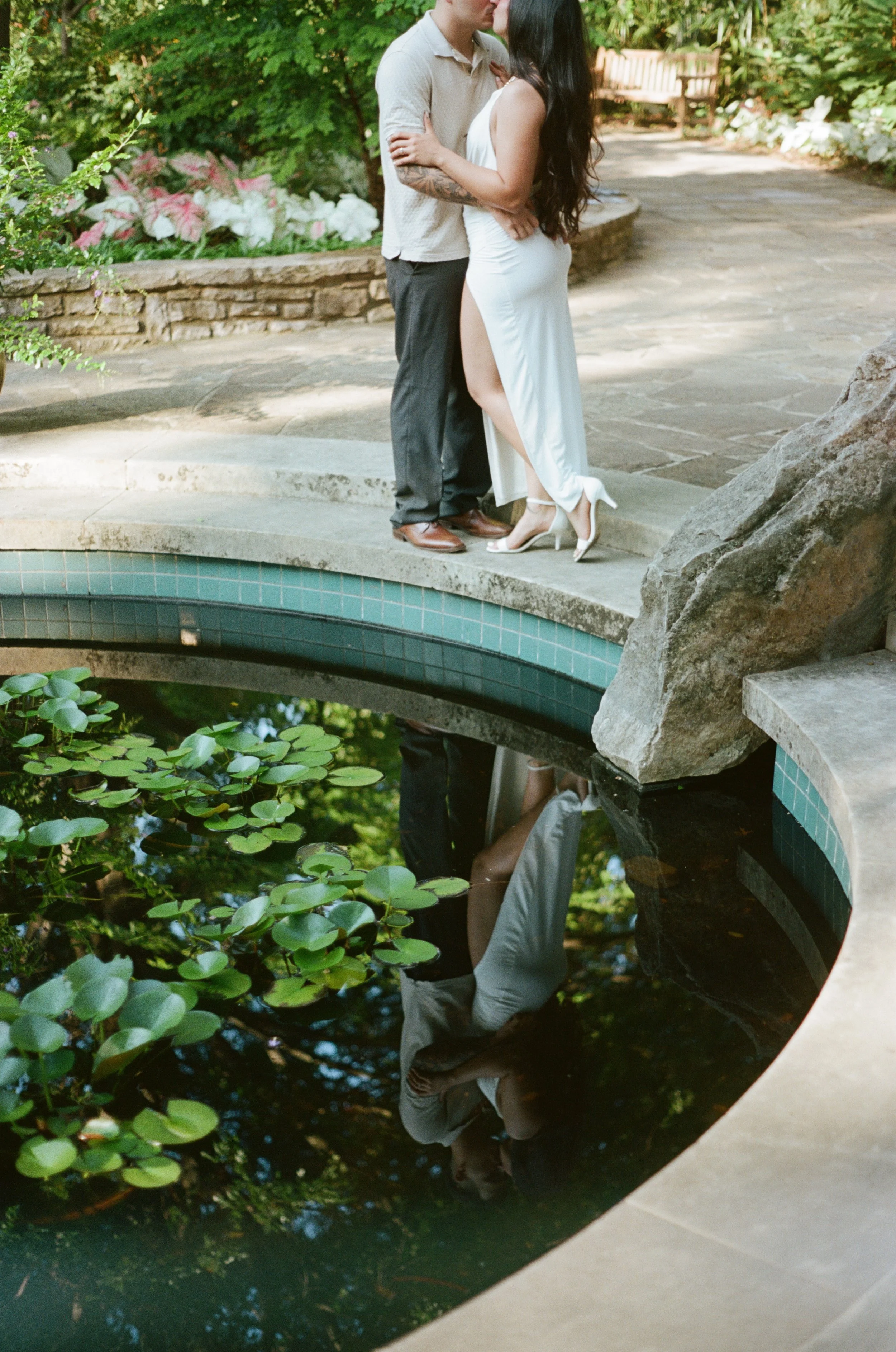 A couple kissing on a small stone bridge over a pond with lily pads, surrounded by greenery and flowers in a garden.