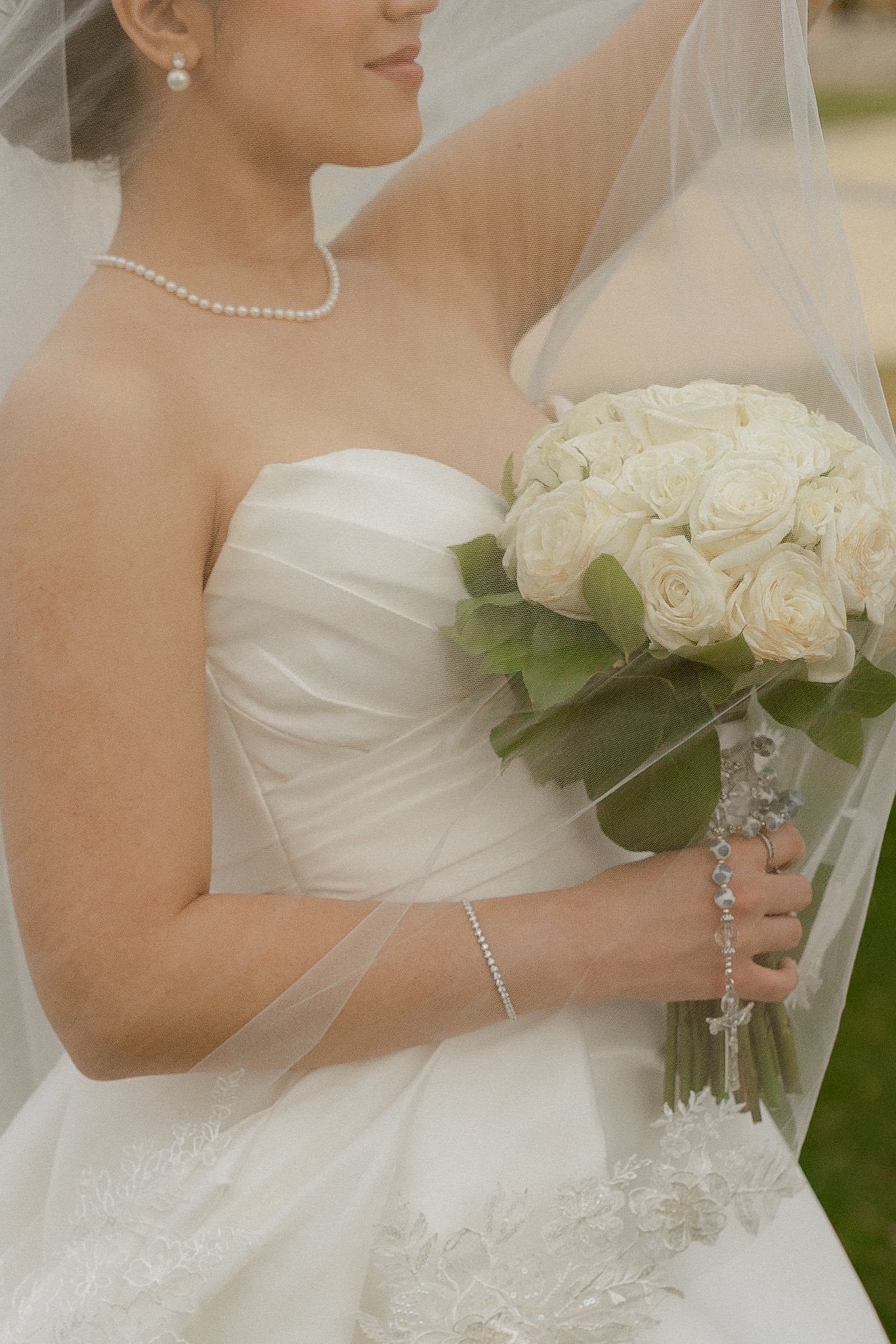 Close-up of a bride wearing a strapless wedding dress, holding a bouquet of white roses, with a sheer veil, pearl necklace, earrings, and bracelet.
