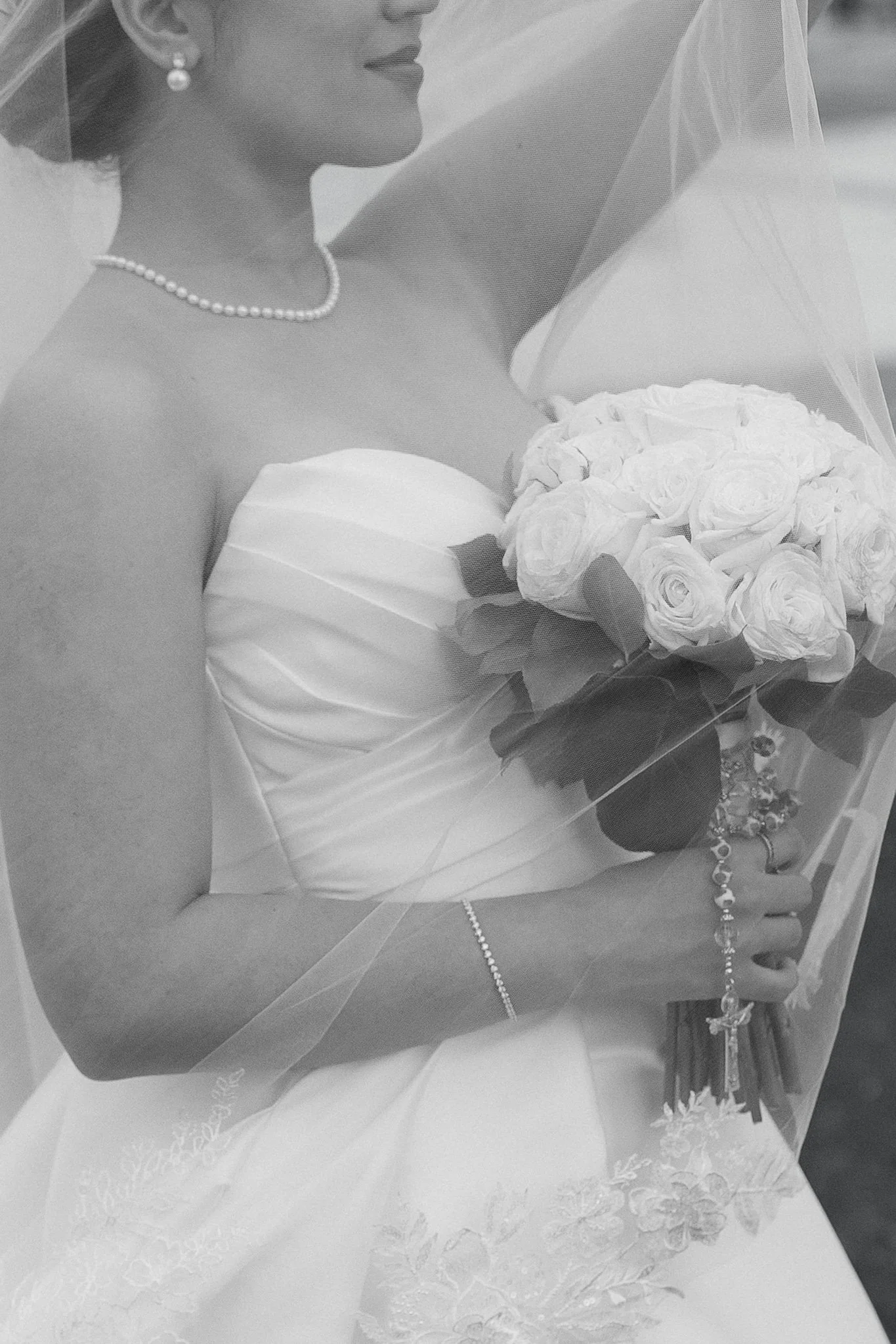 A bride wearing a strapless wedding gown with lace details, pearl necklace, earrings, bracelet, and veil, holding a bouquet of roses and other flowers, with her hand resting on a glass surface.