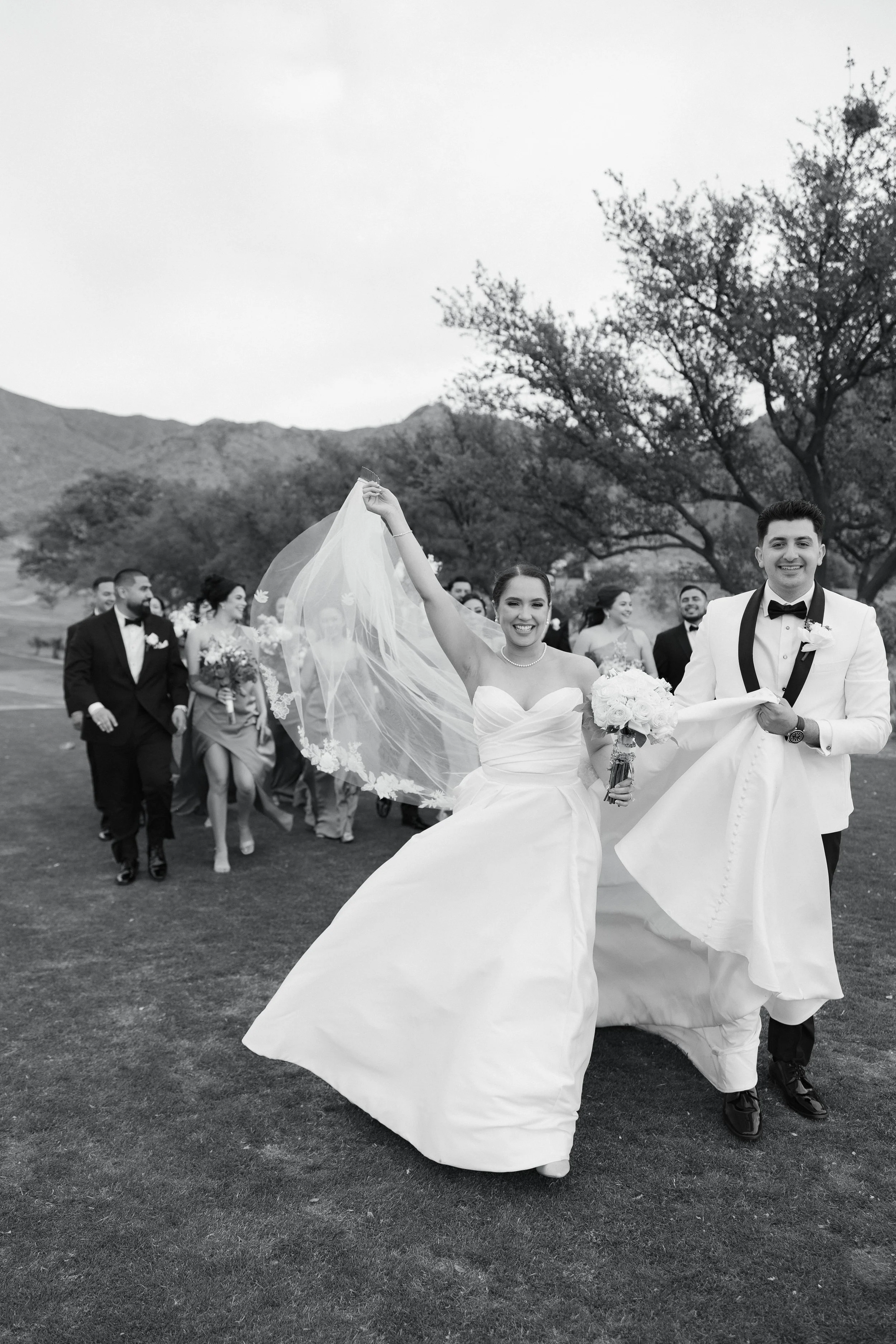 A wedding celebration outdoors with a bride and groom leading a procession of bridesmaids and groomsmen, black and white photo, trees and mountains in the background.