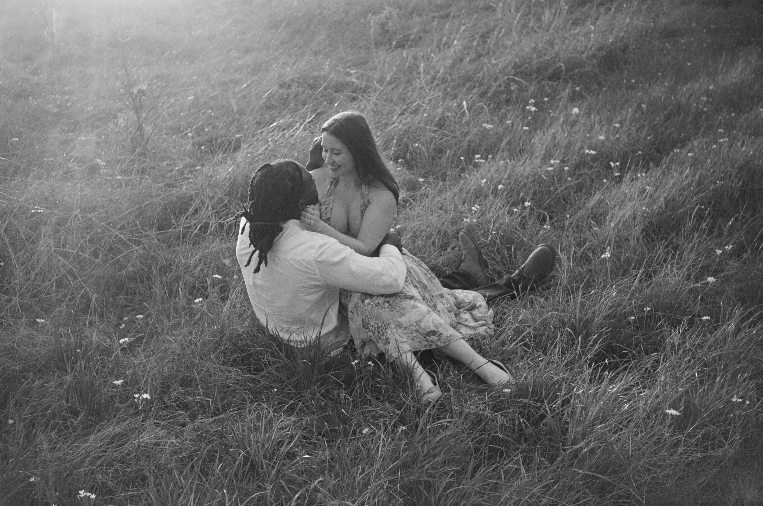 A black and white photo of a couple sitting in a grassy field. The woman is smiling and looking at the man, who is sitting with his legs extended, holding her face and smiling back. The scene is serene with tall grass and small flowers.