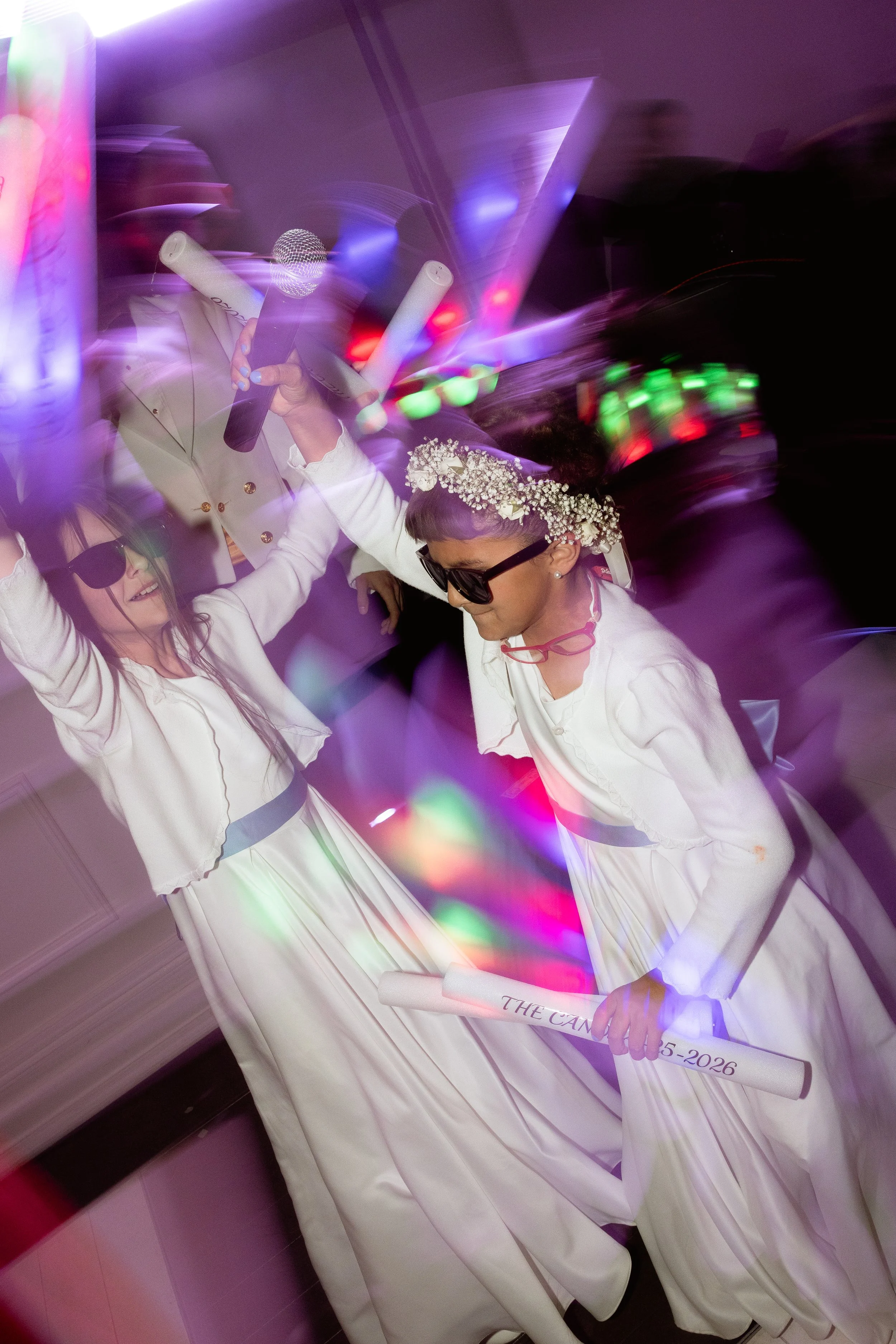 People celebrating at a graduation party, wearing white robes and caps, with some holding scrolls and a microphone, colorful lights in the background.