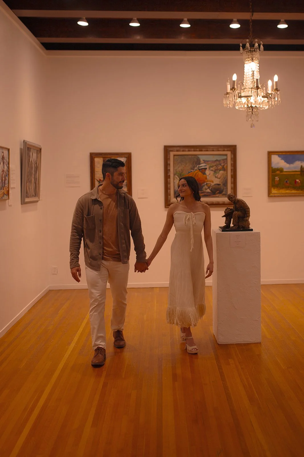 A couple walking hand in hand through an art gallery, holding hands and smiling at each other, with paintings on the walls and a chandelier hanging from the ceiling.