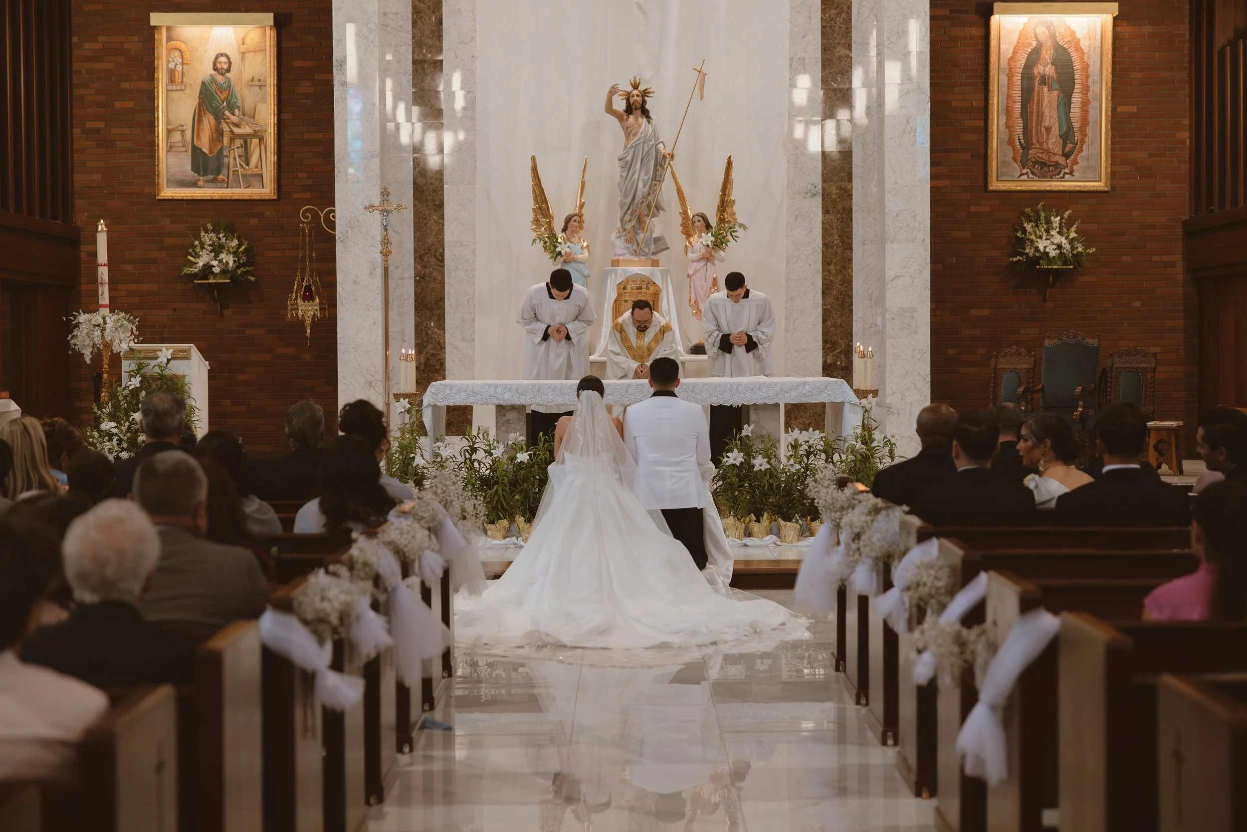 A wedding ceremony in a church with the bride and groom kneeling before the altar. Priests and altar servers are standing behind them. The altar features religious statues and symbols, including Jesus Christ and angels, with floral decorations and li