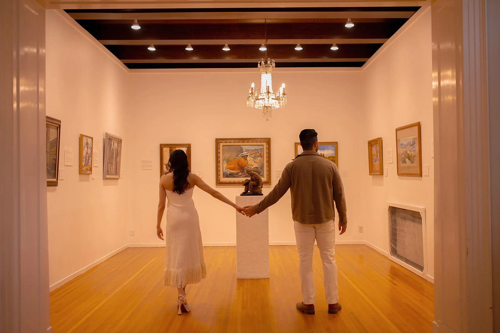 A man and a woman holding hands in an art gallery, viewed from behind. The gallery features paintings on white walls, a chandelier hanging from the ceiling, and a wooden floor.