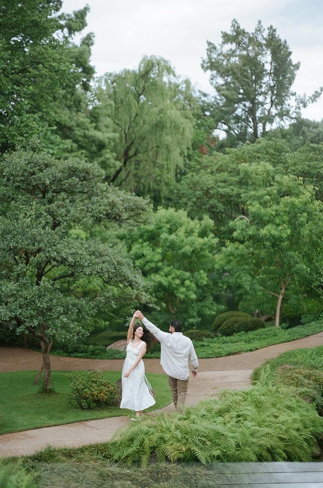 A couple dancing in a lush green park with winding paths and dense trees.