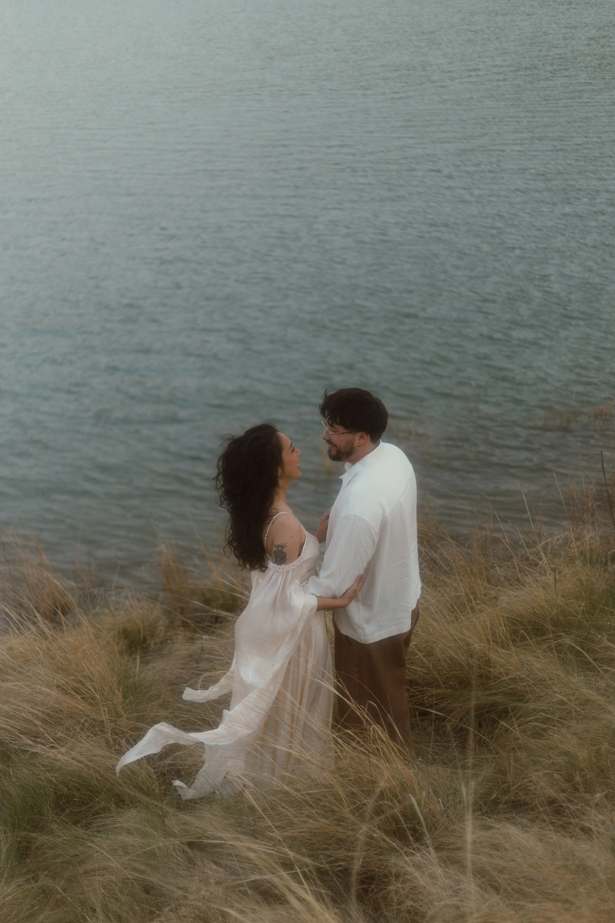 A couple stands on a grassy cliffside near water, looking at each other lovingly.