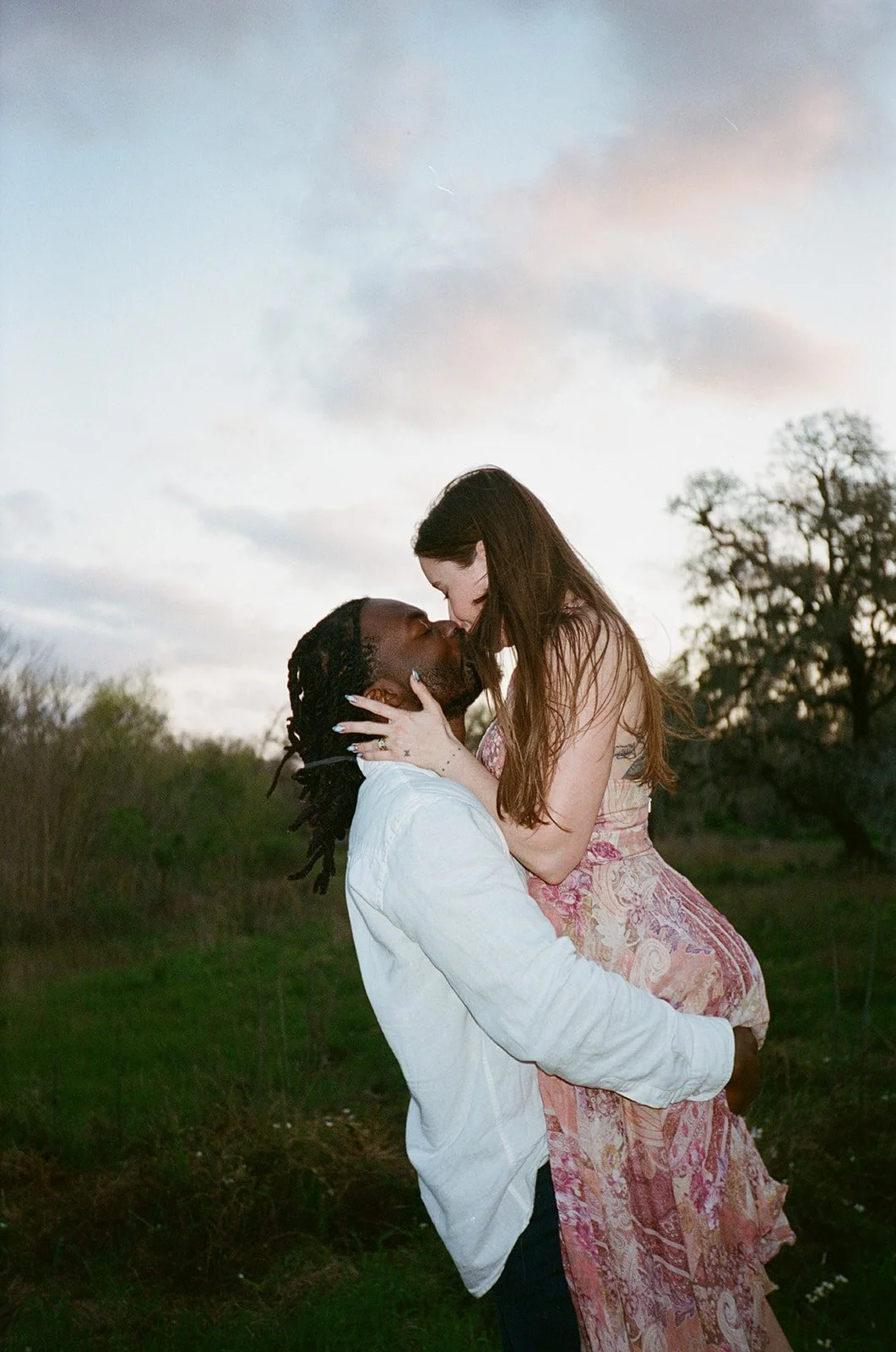 A couple kissing outdoors during sunset, with trees and sky in the background.