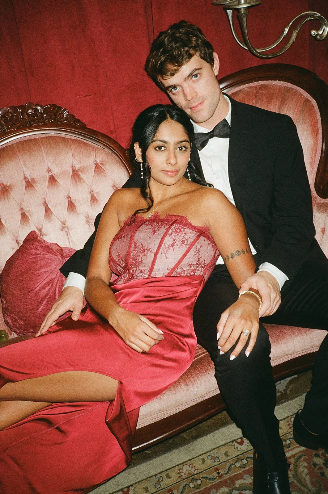 A young man and woman dressed in formal attire sitting on a vintage pink velvet sofa with a red wall background. The woman is wearing a red lace dress and the man is in a black tuxedo with a bow tie.