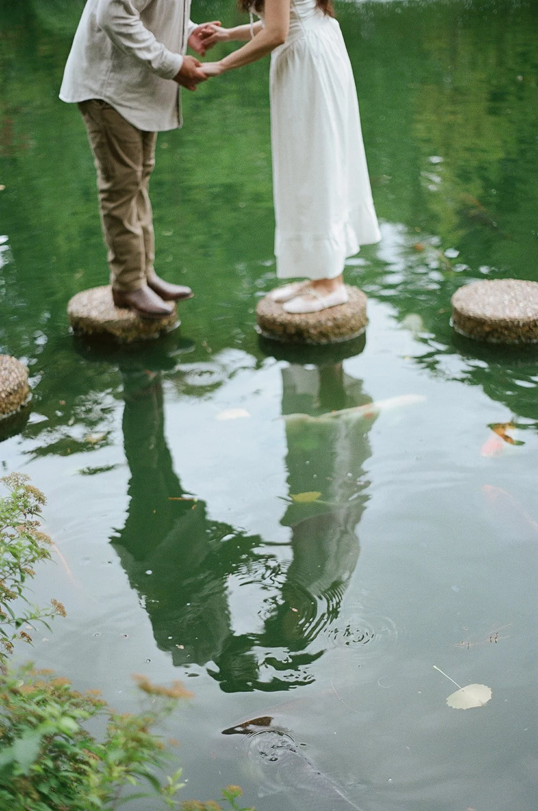 A couple holds hands while standing on circular stepping stones in a pond. The woman wears a white dress, and the man wears tan pants and a light-colored shirt. Fish swim under the stones, reflected in the water.