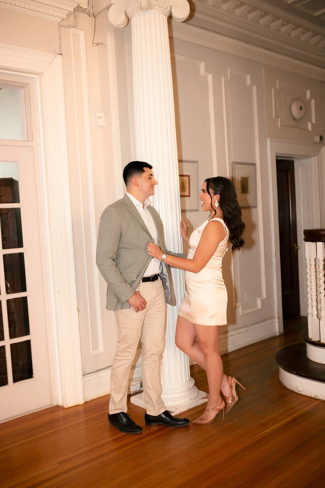 A man in a light gray suit and a woman in a cream-colored dress and high heels touch each other and smile in a formal, elegant room with white walls, a tall white column, and wooden floors.