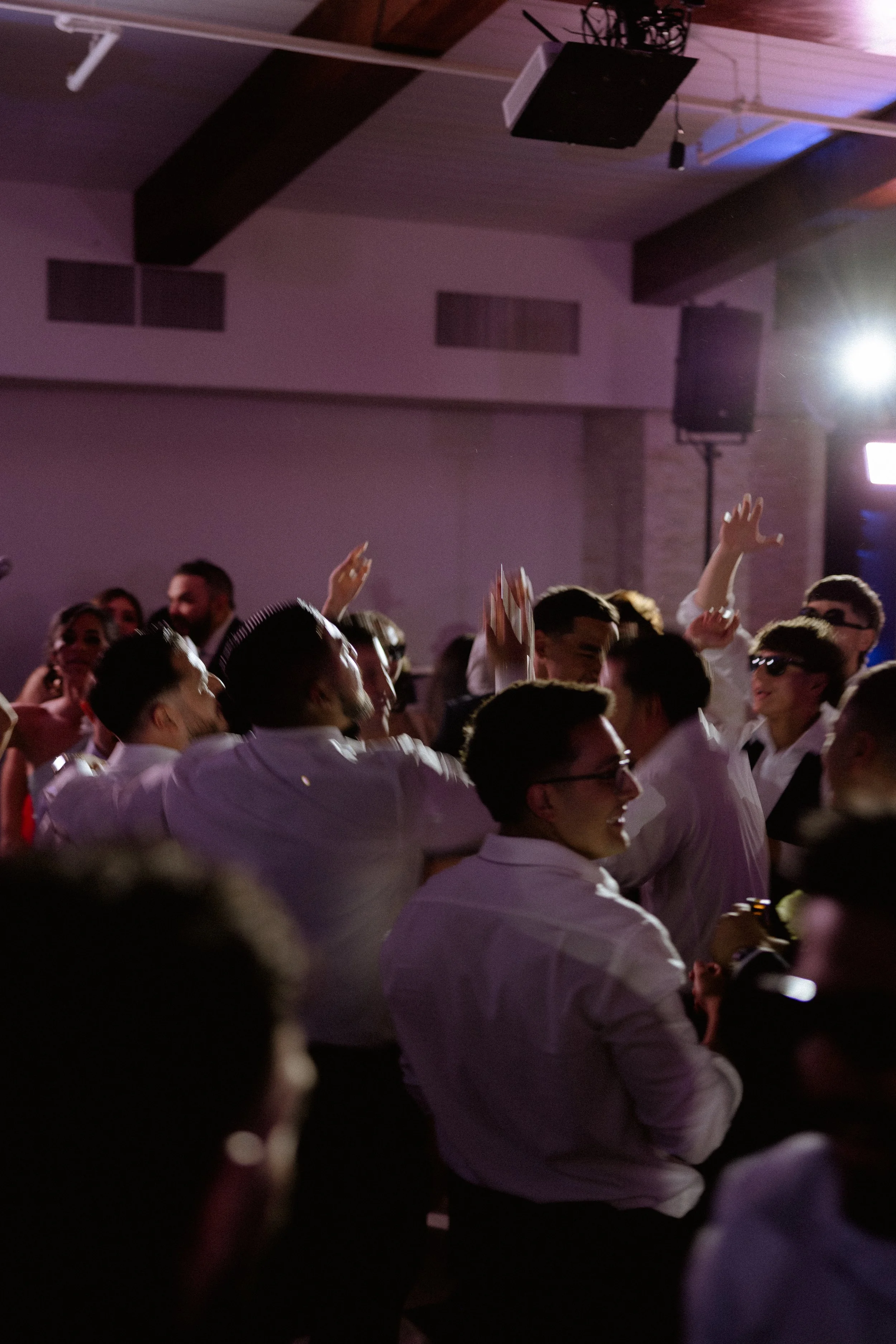Crowd of people gathered at an event with raised hands, wearing white shirts, under dim lighting with some purple and blue highlights, indoors.