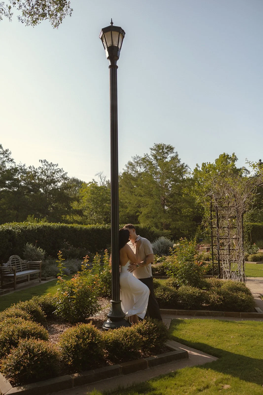 A couple is kissing behind a streetlamp in a lush garden during sunset.