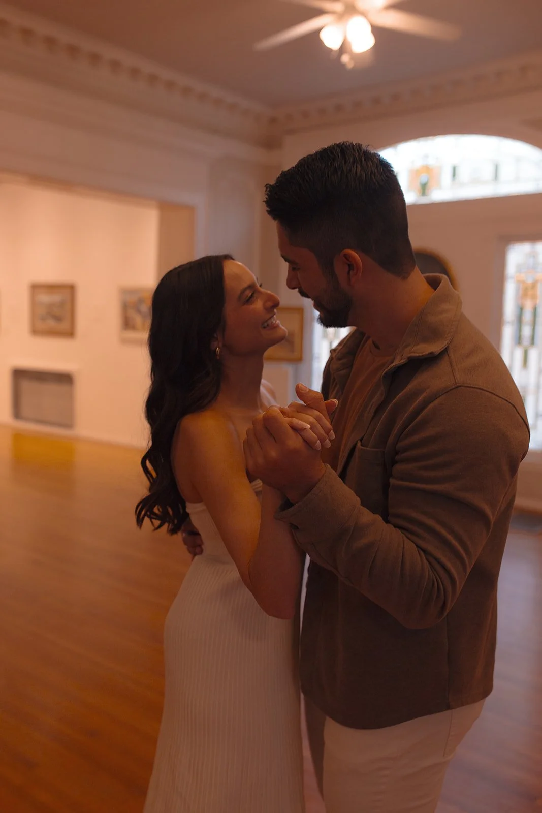 A couple dancing closely, smiling, in a warmly lit room with artwork on the walls and wooden floors.