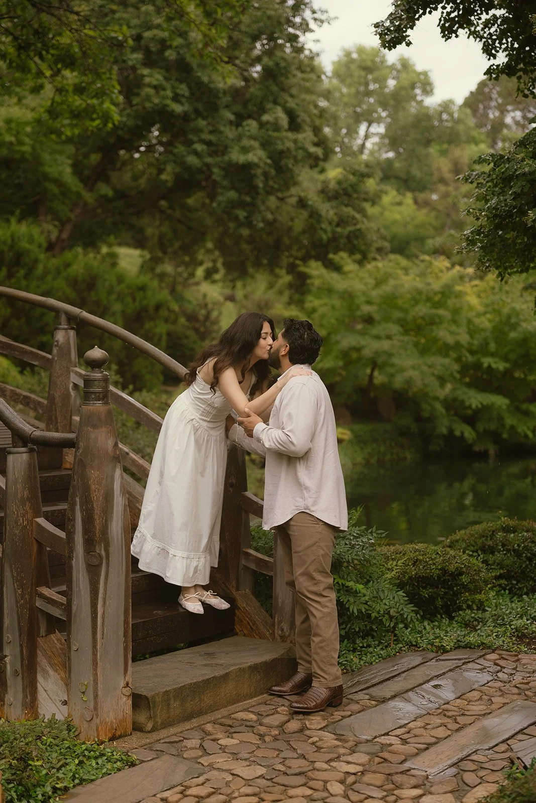 A couple sharing a kiss on a wooden staircase outdoors surrounded by lush greenery and trees.