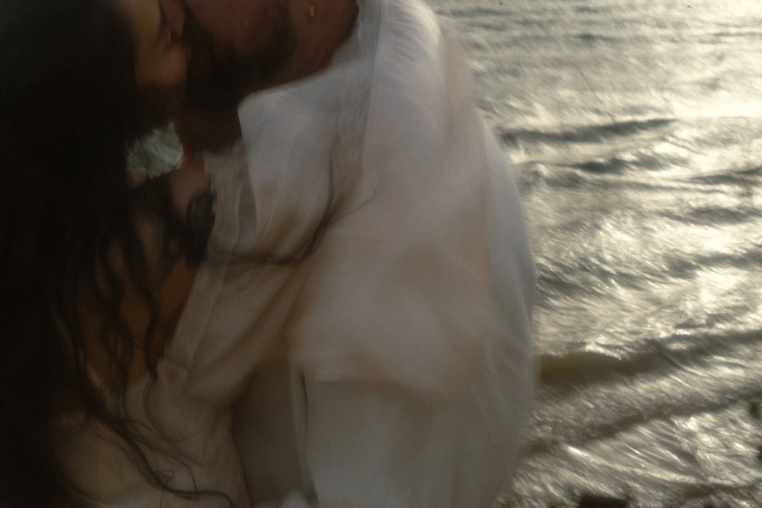 A person with long, dark, curly hair lying down on a bed with white sheets near a textured wall, with sunlight reflecting on the surface.