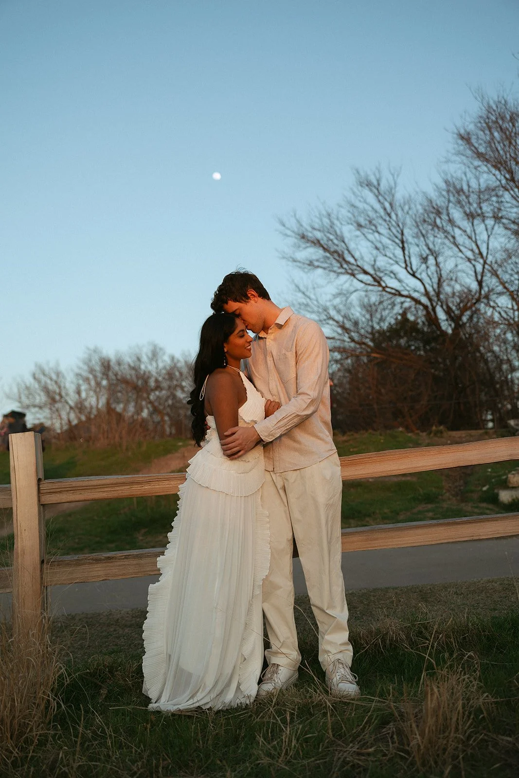 A couple embraces outdoors during dusk, with bare trees and a visible moon in the sky.