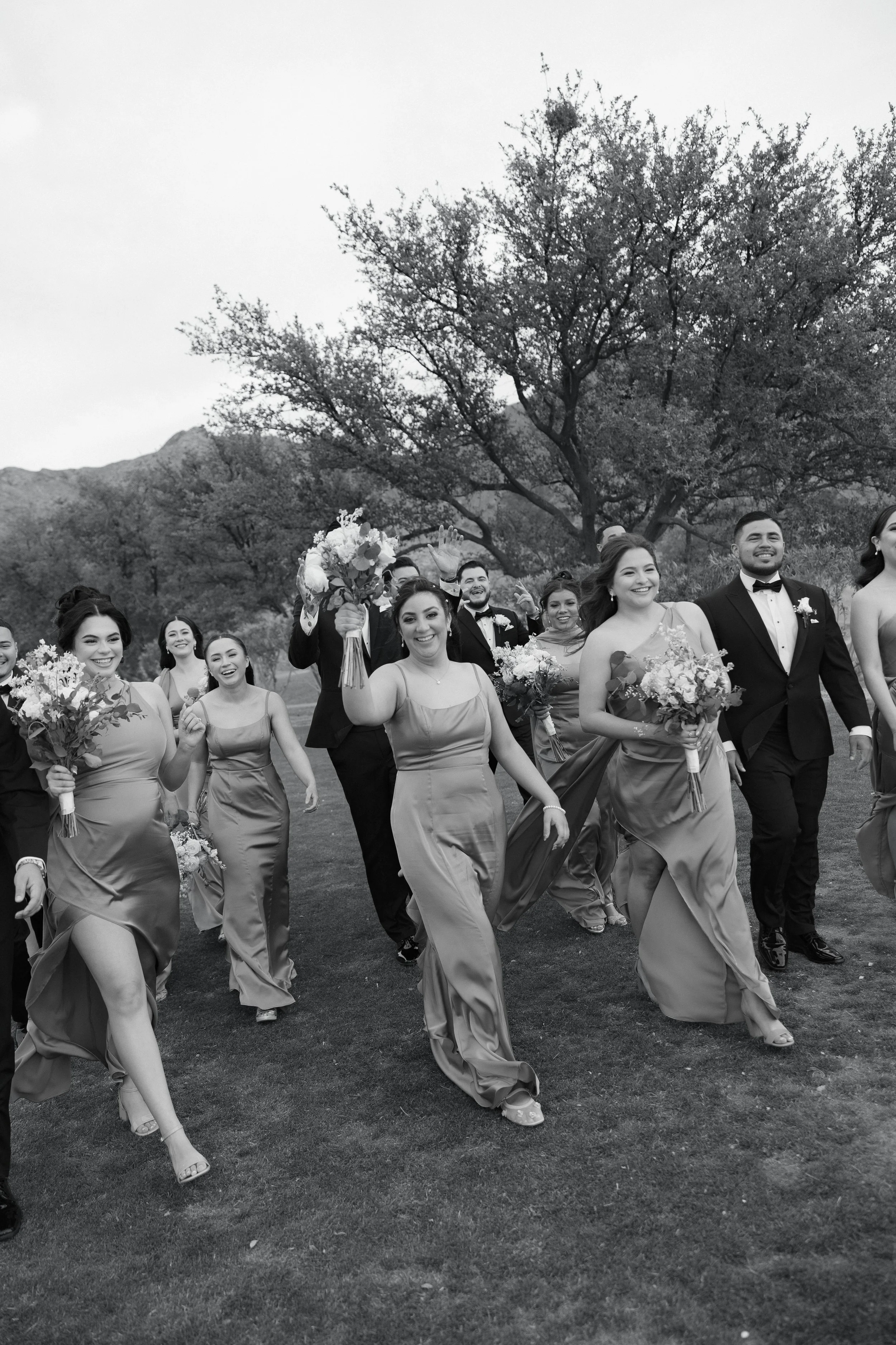 Black and white photograph of a wedding celebration with bridesmaids in matching dresses, groomsmen in tuxedos, holding bouquets, walking outdoors on grass, with a large tree and mountain in the background, and the bride waving to the camera.