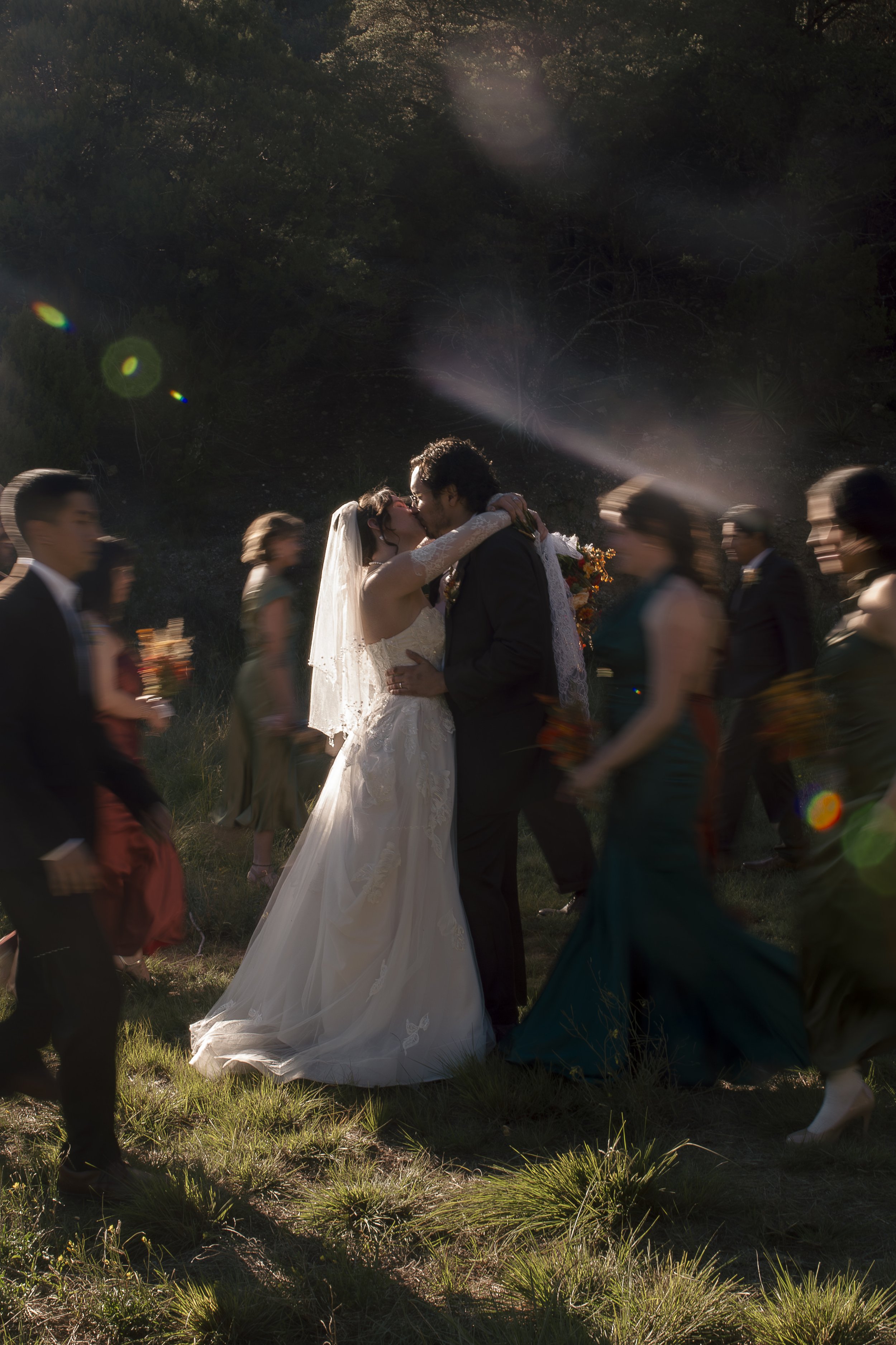 A bride and groom share a kiss during their outdoor wedding ceremony, surrounded by walking guests and greenery, with sunlight filtering through trees.