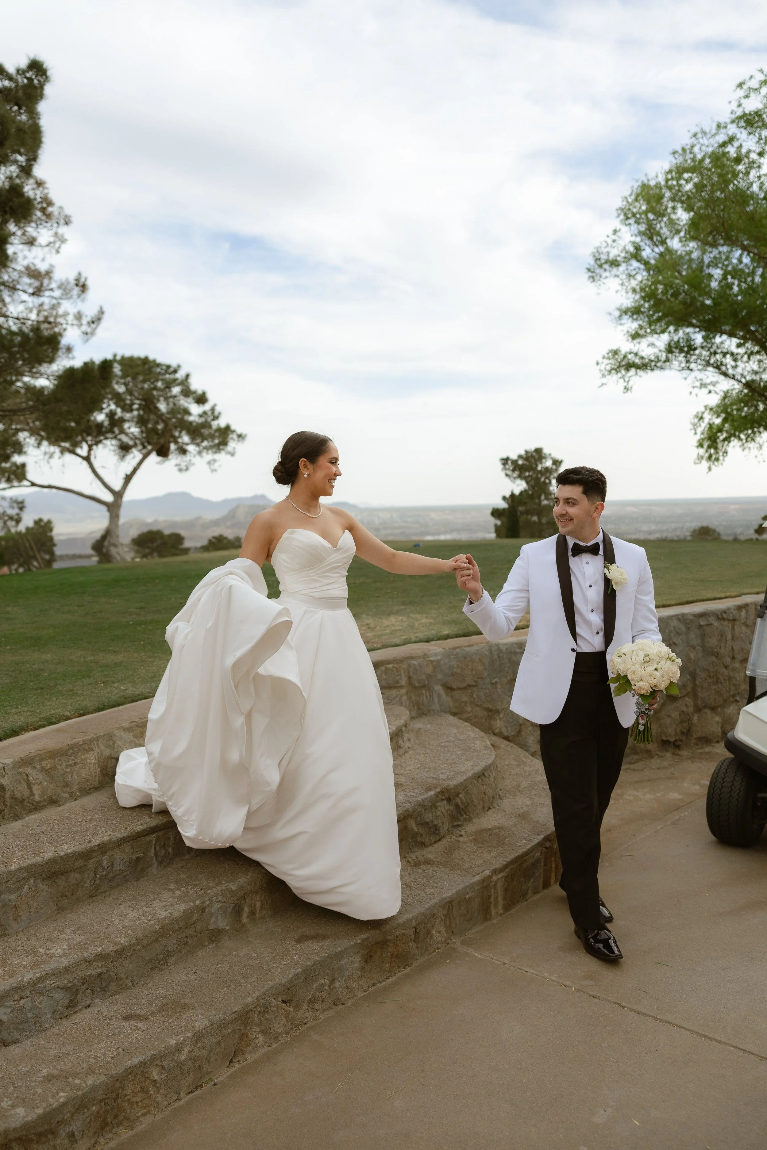 A bride and groom holding hands outdoors during a wedding, with the bride in a white wedding dress and the groom in a tuxedo, smiling at each other with trees and a scenic landscape in the background.