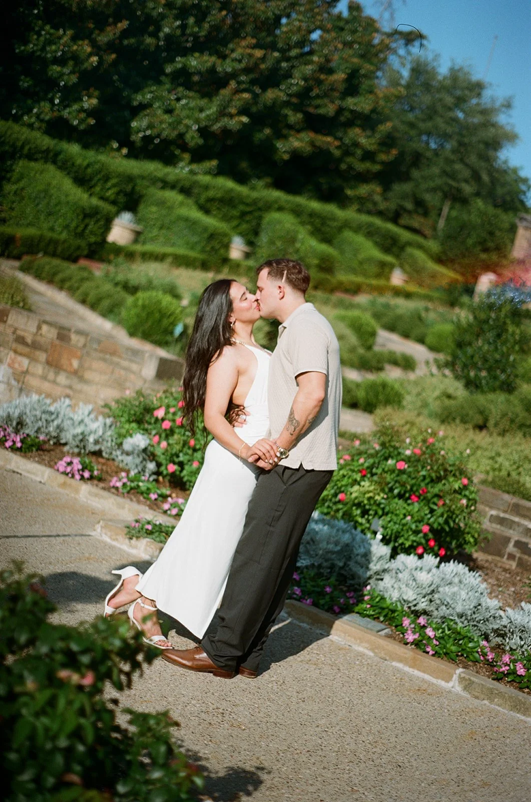 A couple sharing a kiss outdoors in a garden with trimmed bushes and blooming flowers.