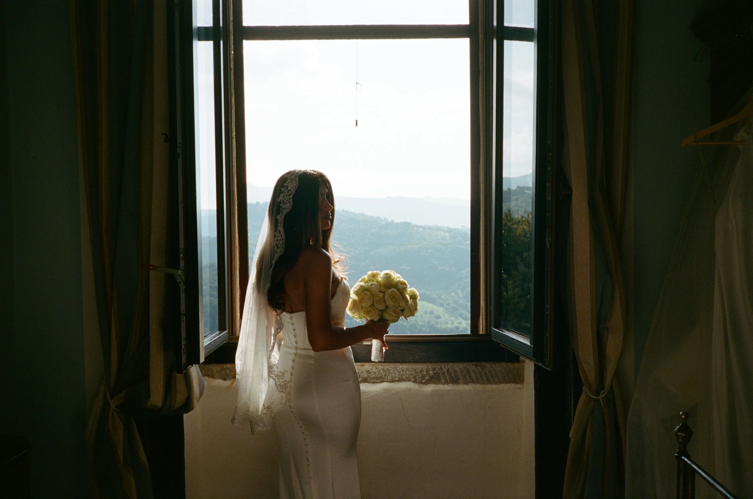 A bride standing in an open window, holding a bouquet of yellow and white flowers, with scenic hills in the background.