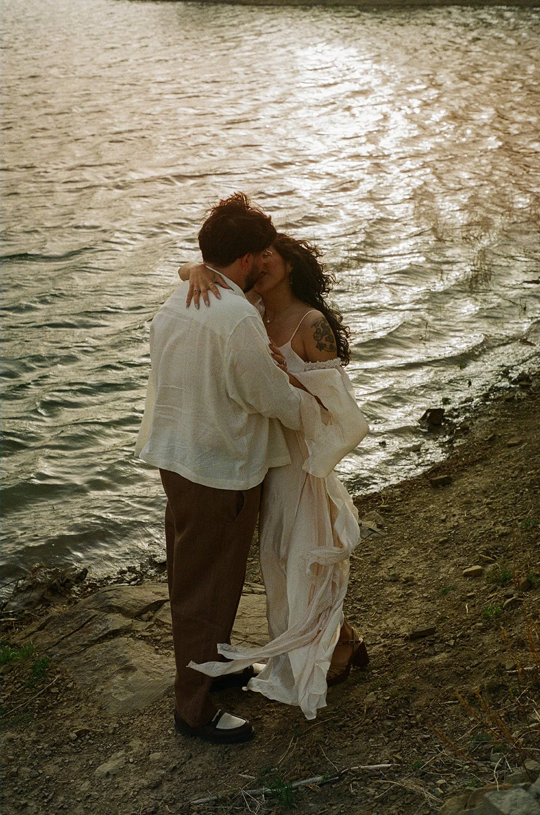 A couple is embracing and kissing by the water's edge during sunset or sunrise.