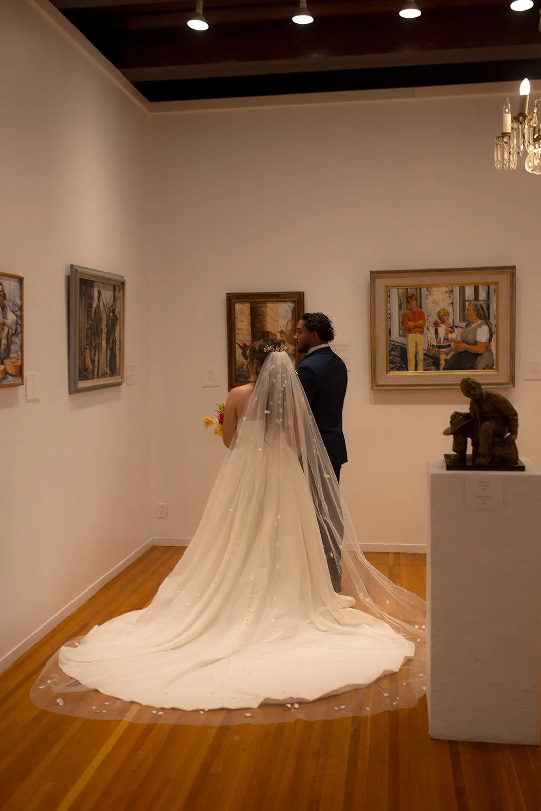 A bride and groom standing together in an art gallery, facing each other, with a bride wearing a long white dress and veil, and a groom in a dark suit. There are paintings on the walls and a sculpture on a pedestal nearby.