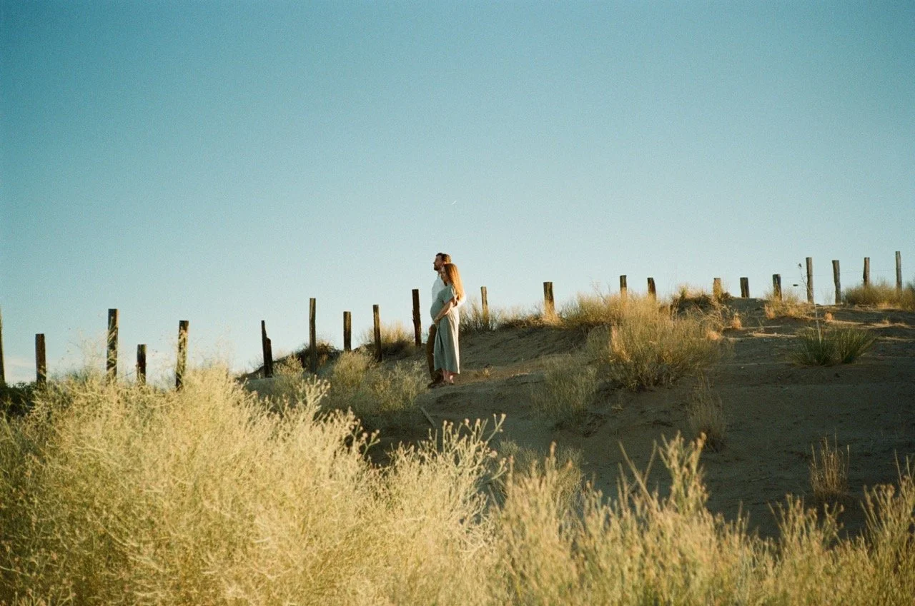 A pregnant woman with long hair walking along a sandy path on a hill, bordered by wooden fence posts and dry bushes, under a clear blue sky.