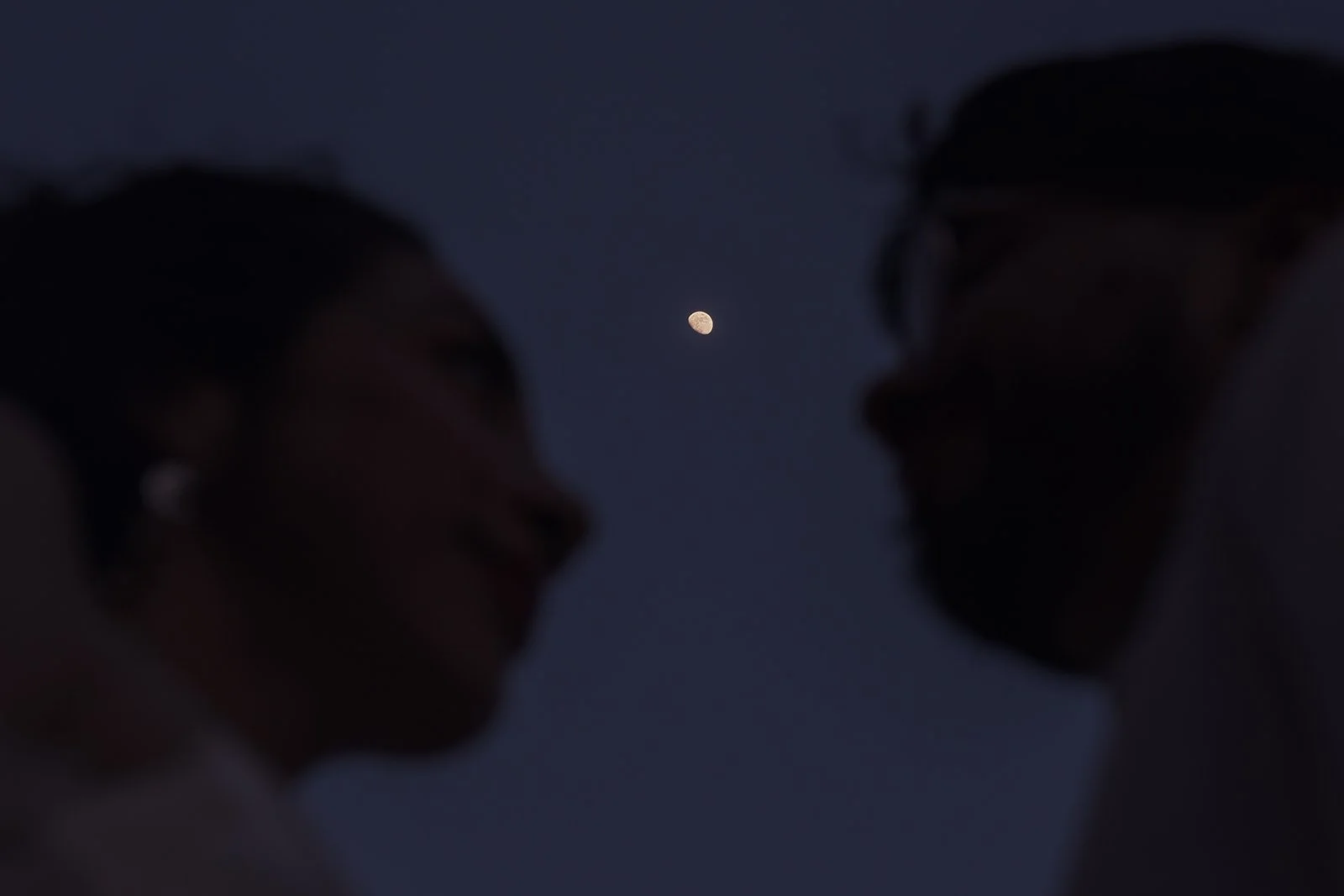 Silhouettes of a couple facing each other at night with the moon visible between them in the dark sky.