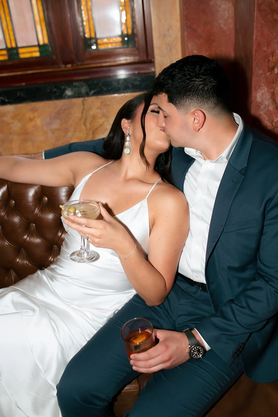 A couple dressed in formal attire sharing a kiss while holding drinks at a bar or lounge.