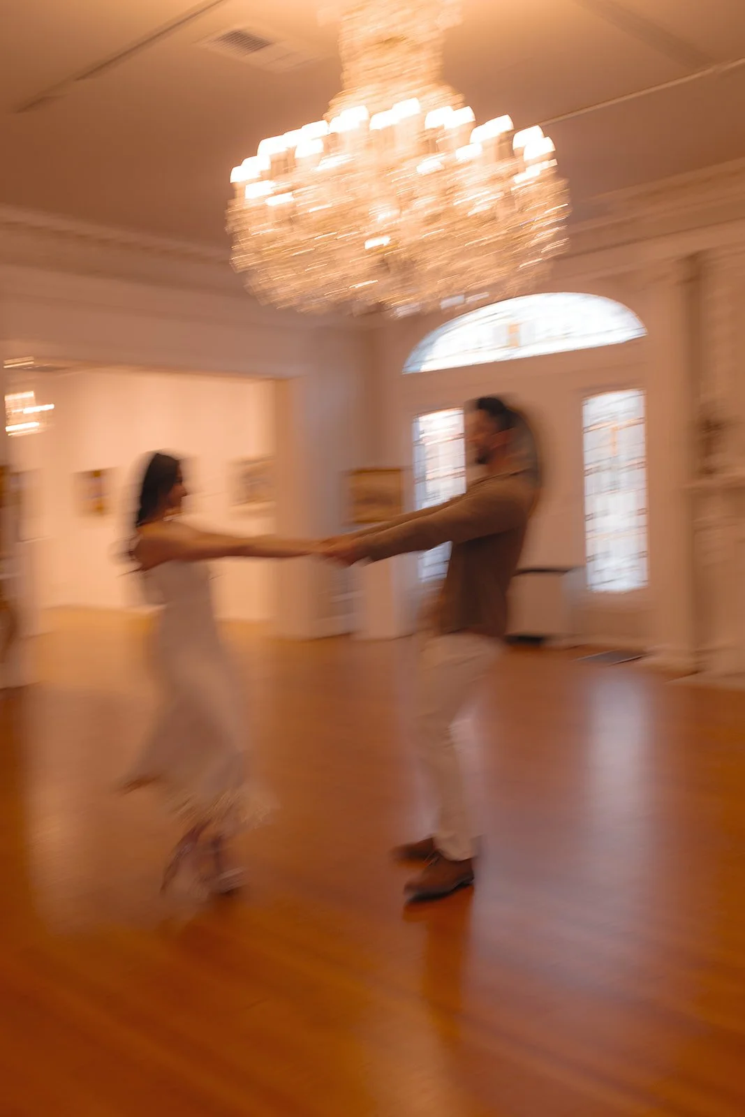 A blurred photo of a couple dancing in a luxurious room with a large chandelier, high windows, and wooden flooring.