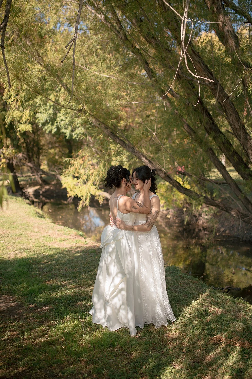 Two women in wedding dresses embracing and smiling outdoors under tree branches near a stream.