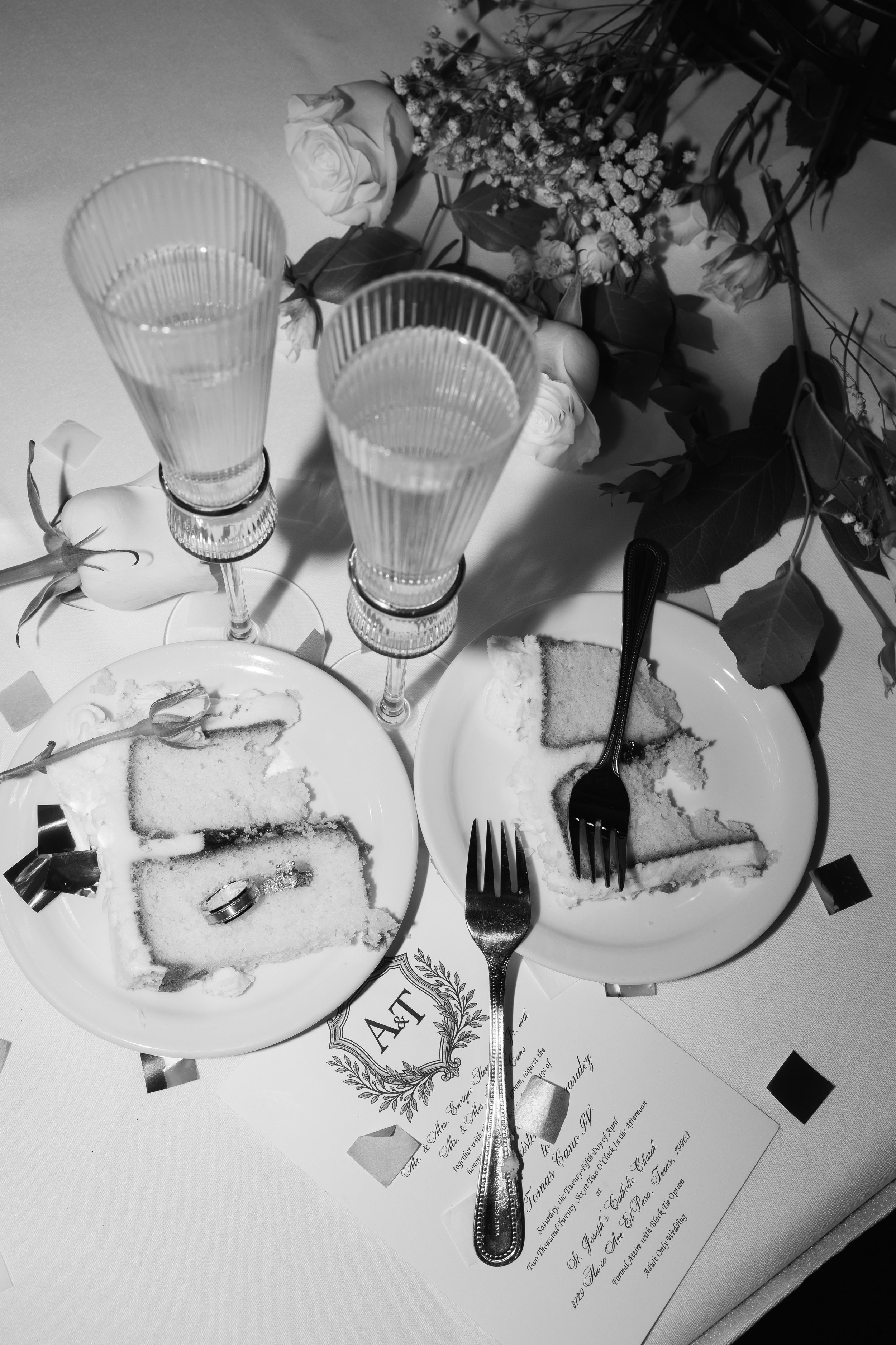 Two plates with partially eaten slices of cake, plastic rings, and utensils; two tall, ribbed glasses with water; a bouquet of flowers with roses and other small flowers on a table with a wedding invitation underneath.