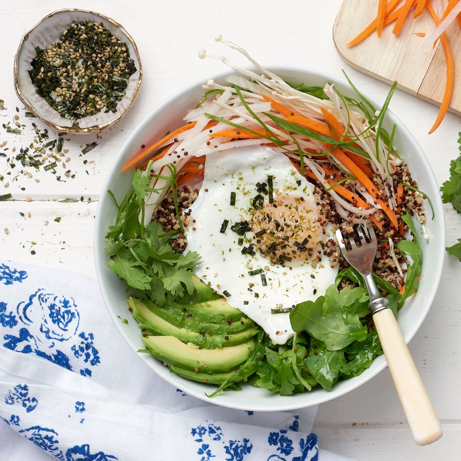 A bowl of salad with sliced avocado, mixed greens, shredded carrots, and a poached egg, topped with chopped herbs and black sesame seeds, on a white wooden surface with a fork and a small dish of herbs and sesame seeds nearby.