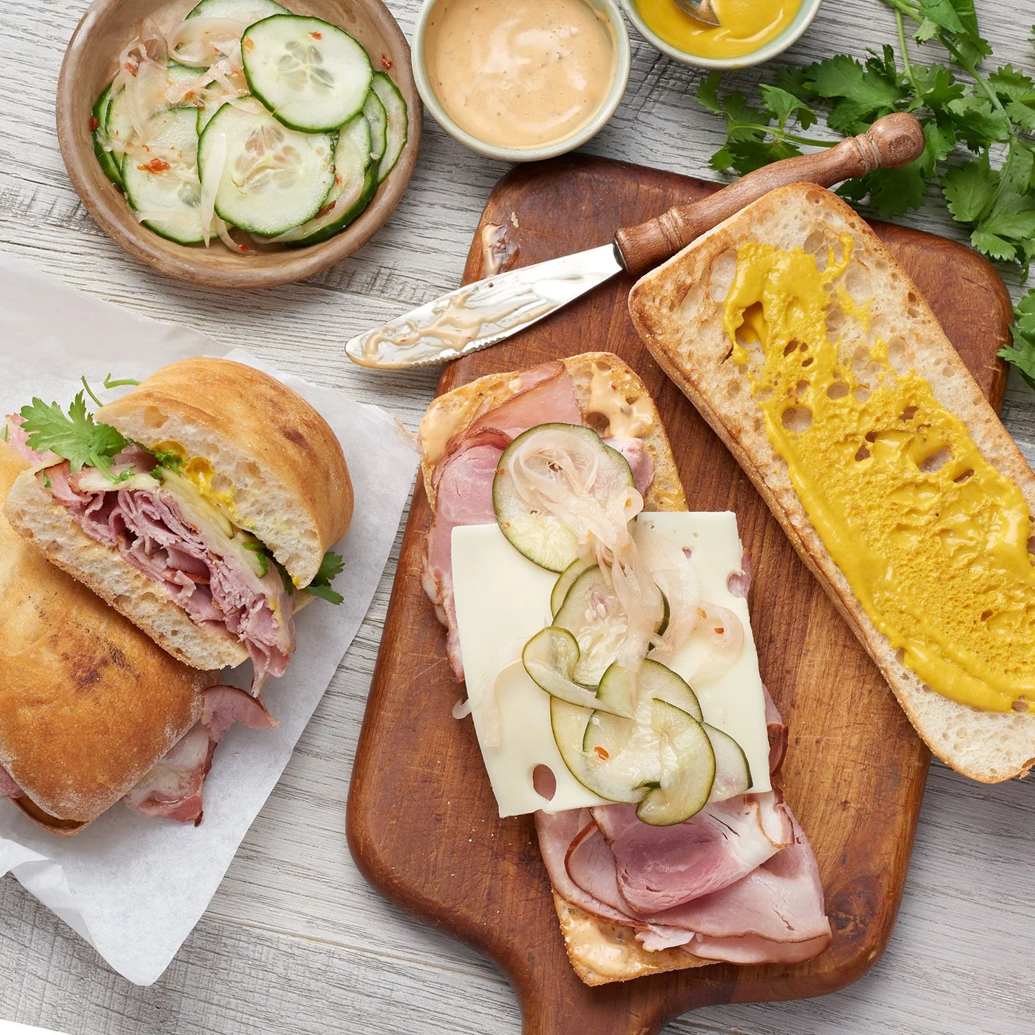 A cuban style sandwich on a wooden cutting board, with cheese and cucumber salad and small bowls of dressing.