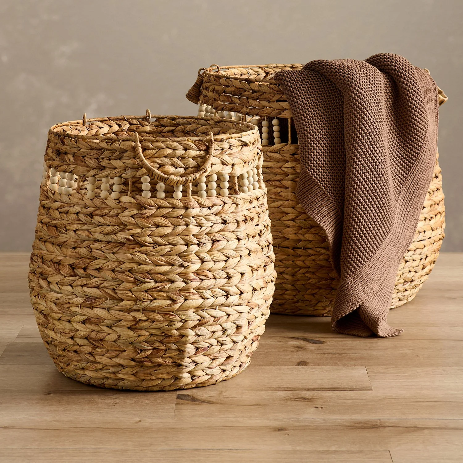 pair of woven storage baskets with brown draping blanket inside set in-front of textured taupe wall and wood floor